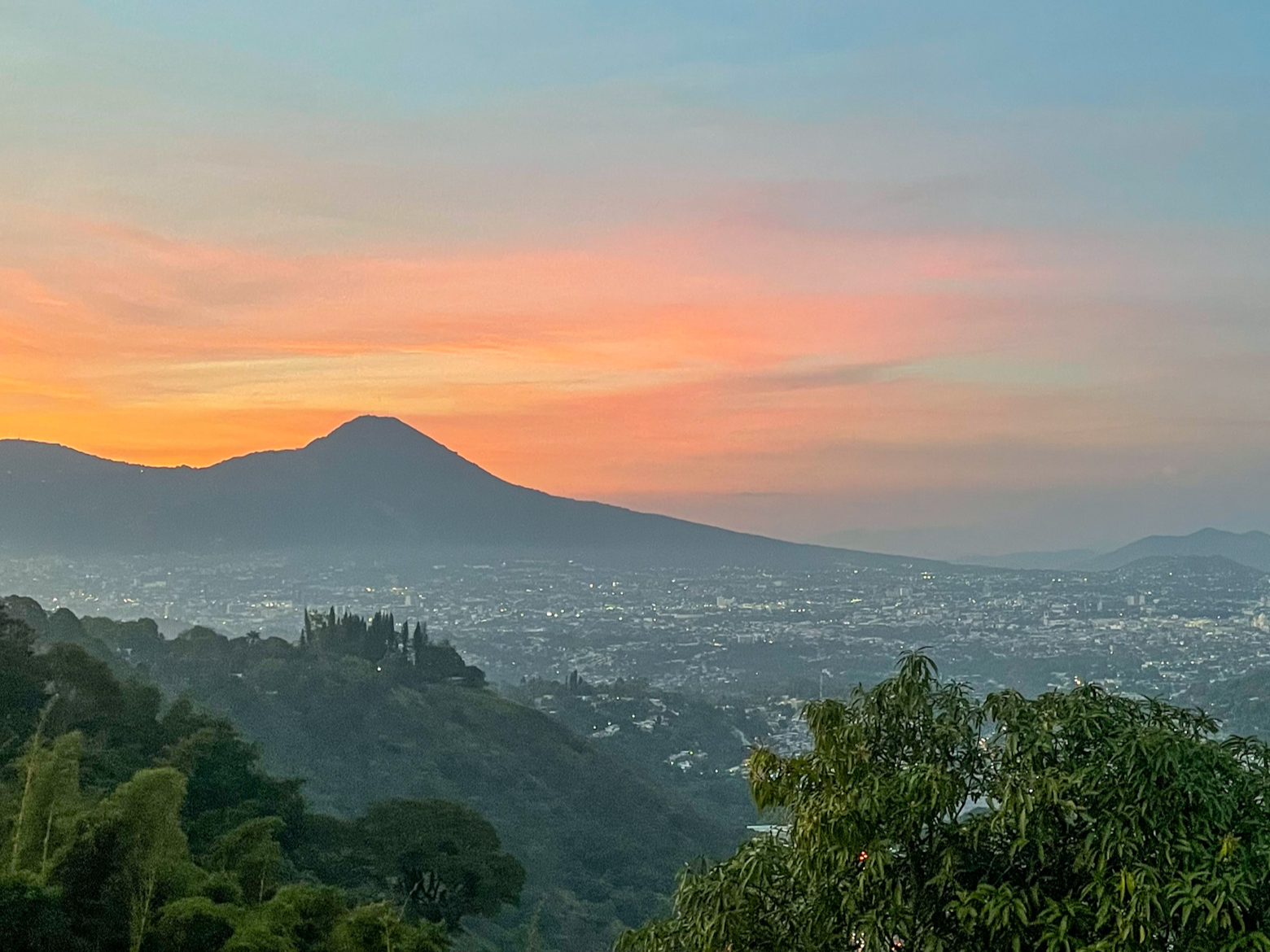 Sunset over a city with a mountain in the background, trees in the foreground, colorful sky with pink and orange hues.