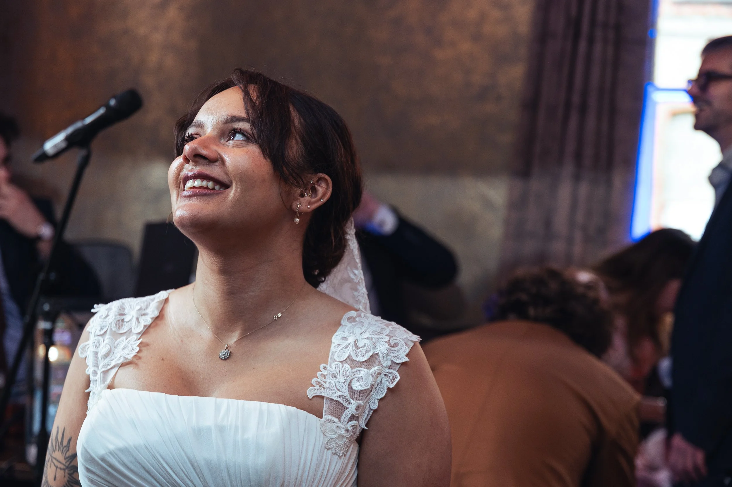 A woman in a white dress with lace shoulders and short dark hair is smiling and looking upward, possibly during a wedding celebration, with a microphone, several people, and a warm indoor setting in the background.