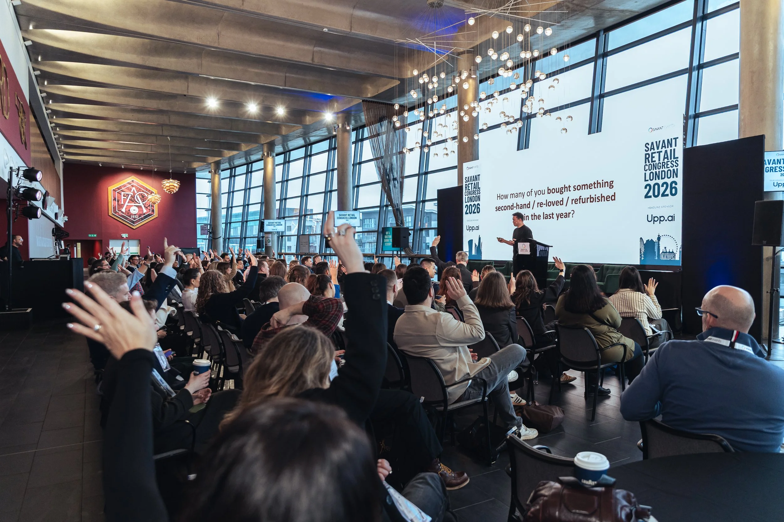 A large audience sitting in a conference room, raising their hands during a presentation. A male presenter stands in front of a large screen displaying information for the Savant Retail Congress London 2026 event. The room has high ceilings, large wi