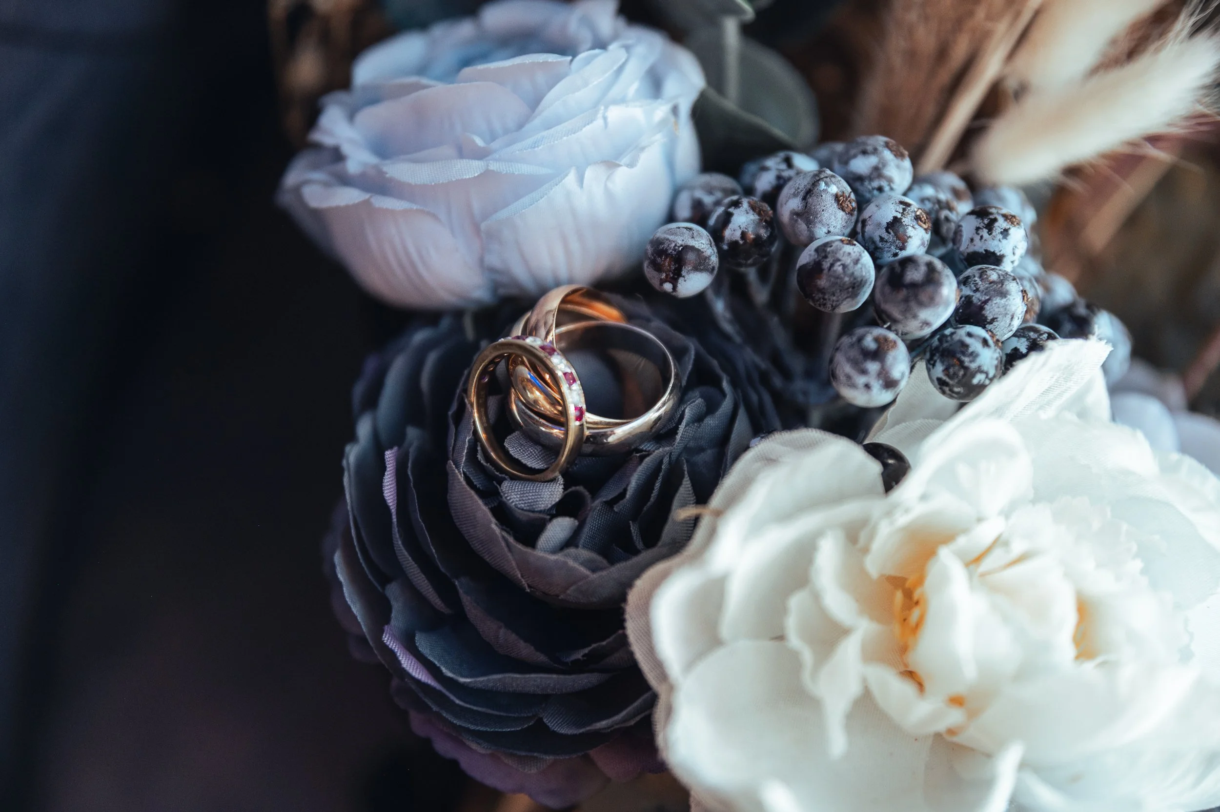 Wedding rings resting on black and gray artificial flowers with white flowers and berries nearby.