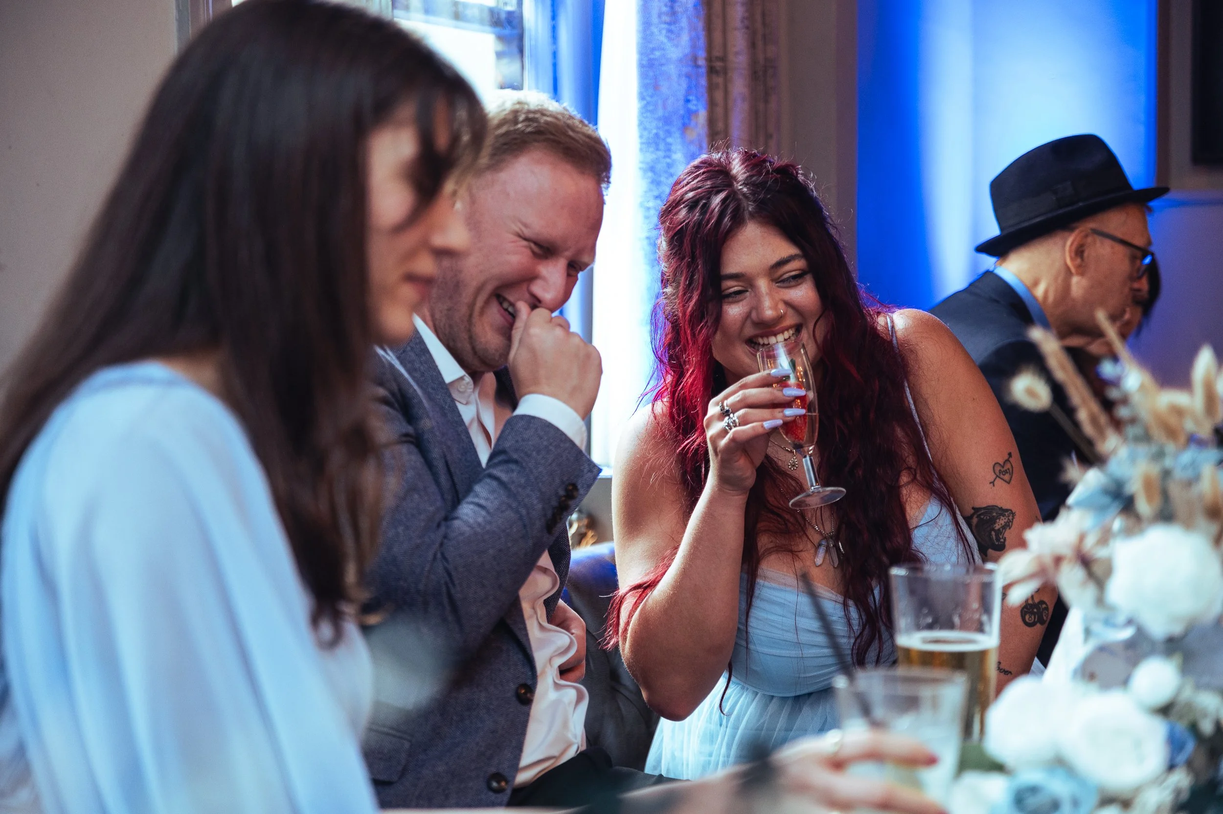 People sitting at a table, smiling and laughing during a celebration, with drinks in hand, in a well-lit room with blue lighting.