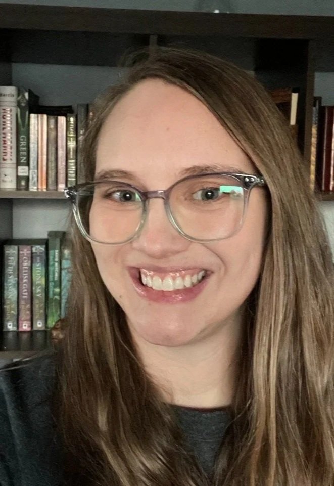 Photo of a smiling, white, blond woman with teal and purple glasses in front of a bookshelf