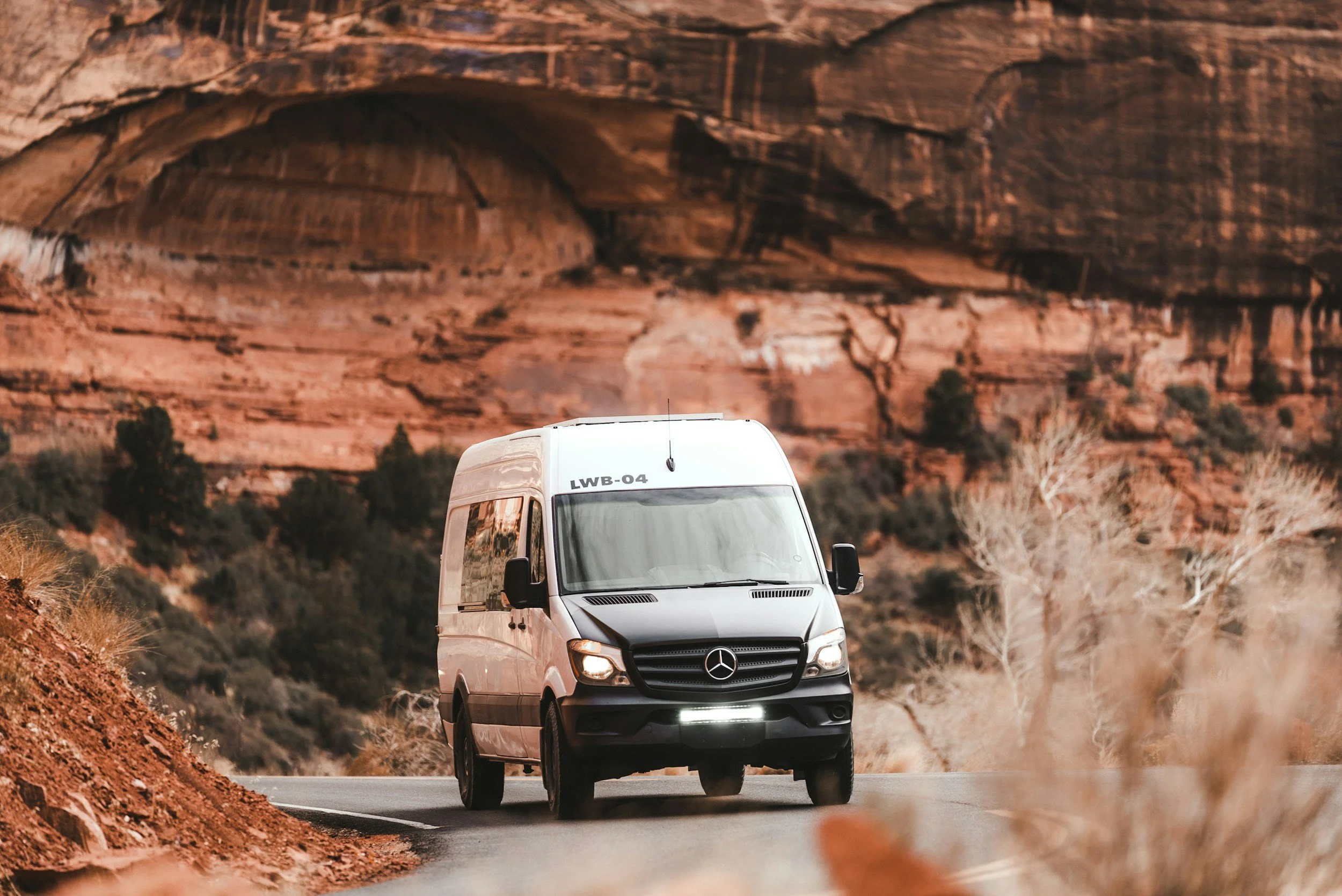 Two people standing in front of a white van with a logo, in a rocky desert landscape with large cliffs in the background. They are smiling, dressed casually for outdoor activity, with bright sunlight and a clear blue sky.