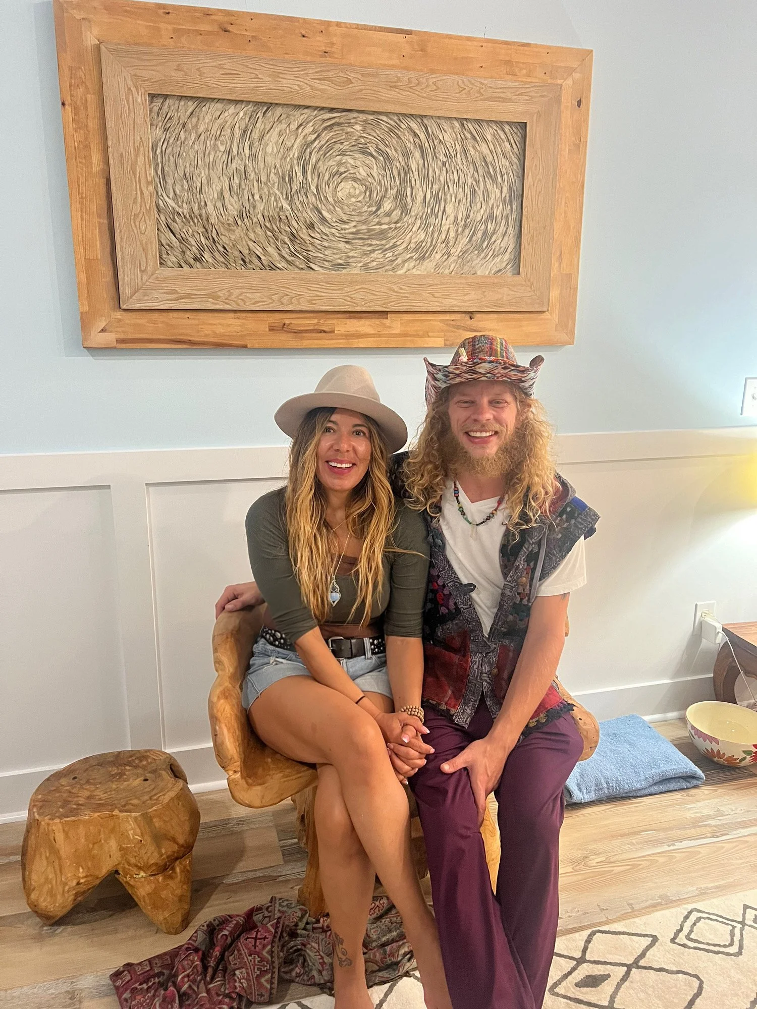 A smiling woman and man sitting together on a wooden chair, holding hands. The woman wears a wide-brimmed hat, a green top, and denim shorts. The man wears a colorful hat, a patterned vest over a white t-shirt, and maroon pants. They are in a cozy room with a large abstract painting on the wall and wooden furniture.