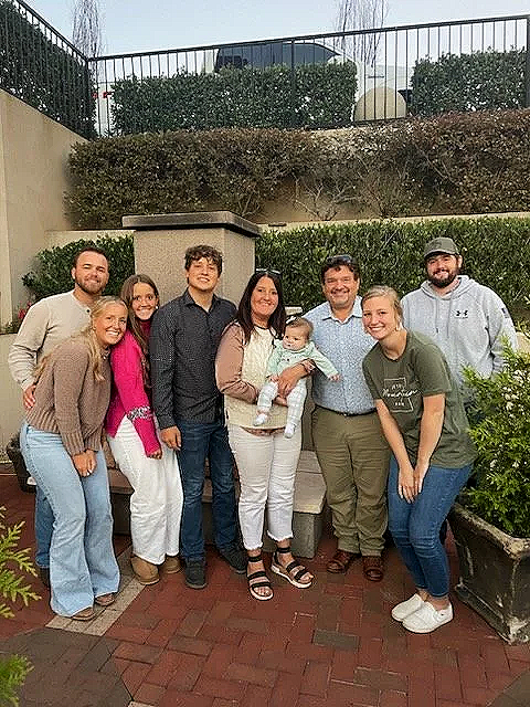 Group of eight adults and one baby outdoors in a garden, posing for a photo. They are standing on red brick paving with green bushes and a low wall behind them.