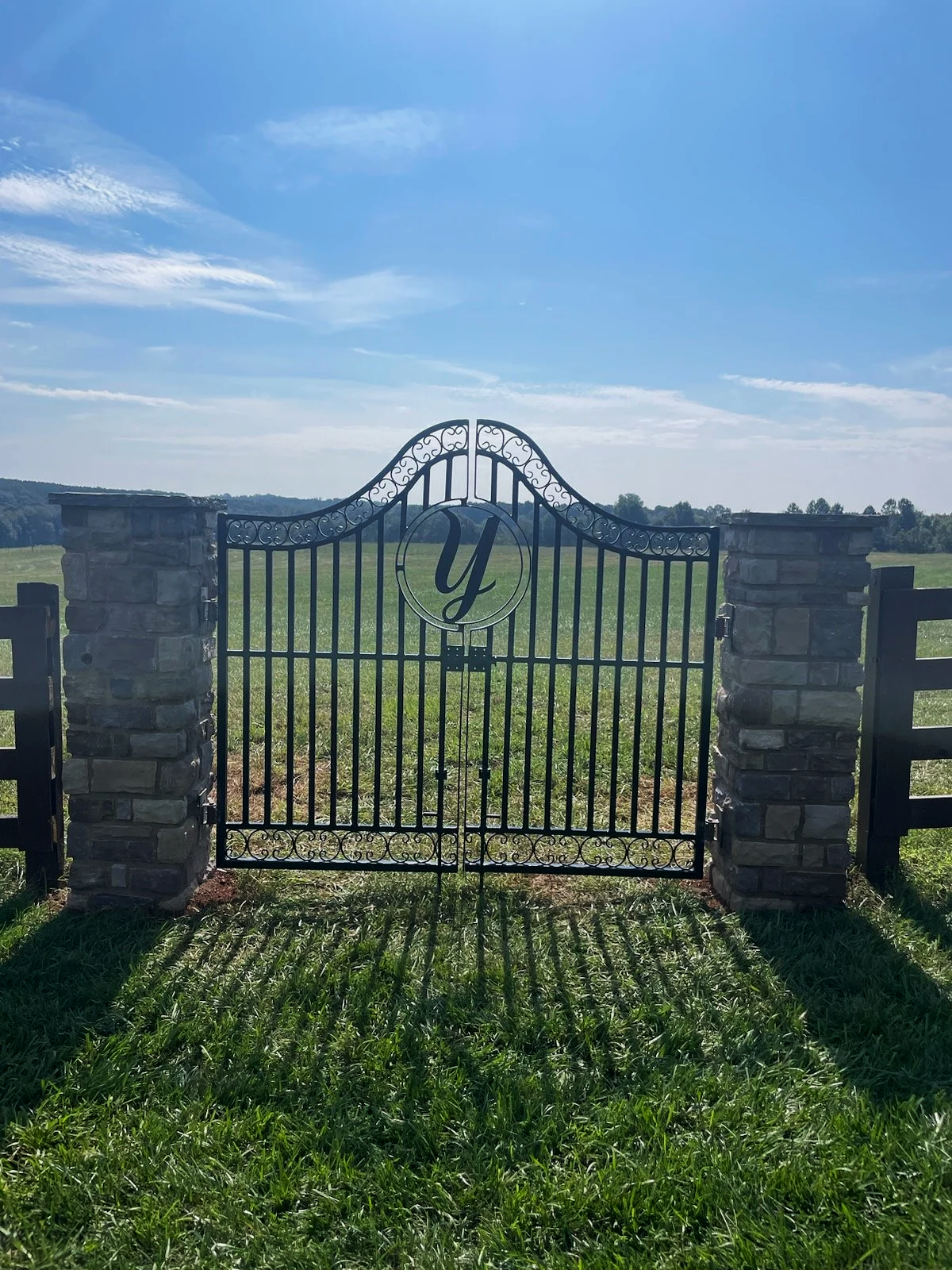 Black wrought iron gate with a circular emblem centered, displaying a letter 'Y', set between two stone pillars, leading to a grassy field with a blue sky and some scattered clouds in the background.