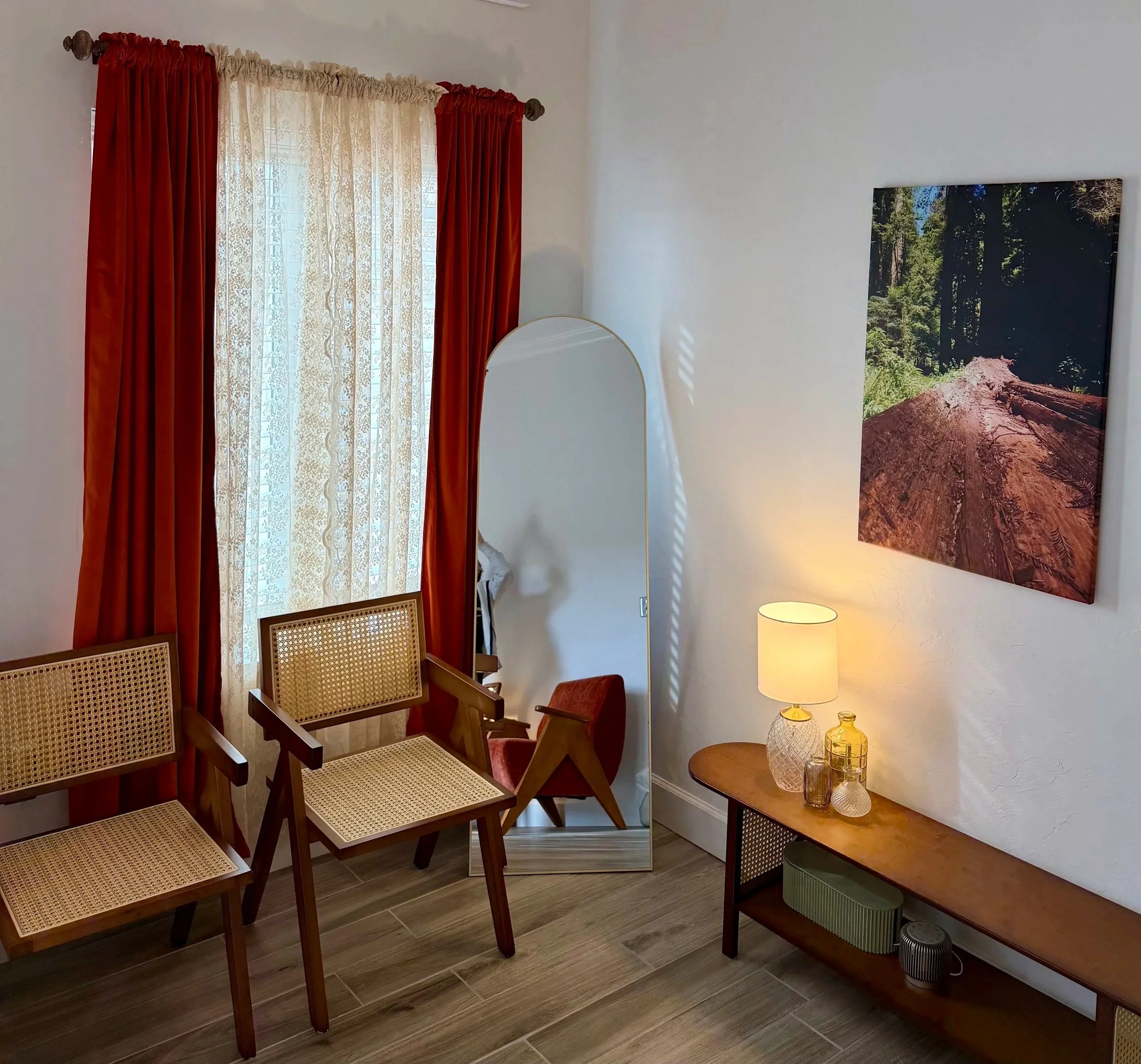 A room with a window with beige lace curtains and orange drapes, two wooden chairs with woven cane seats and backs, a tall mirror, a wooden side table with a lamp and glass jars, a large wall print of a forest and a tree trunk, and a wood-look floor.