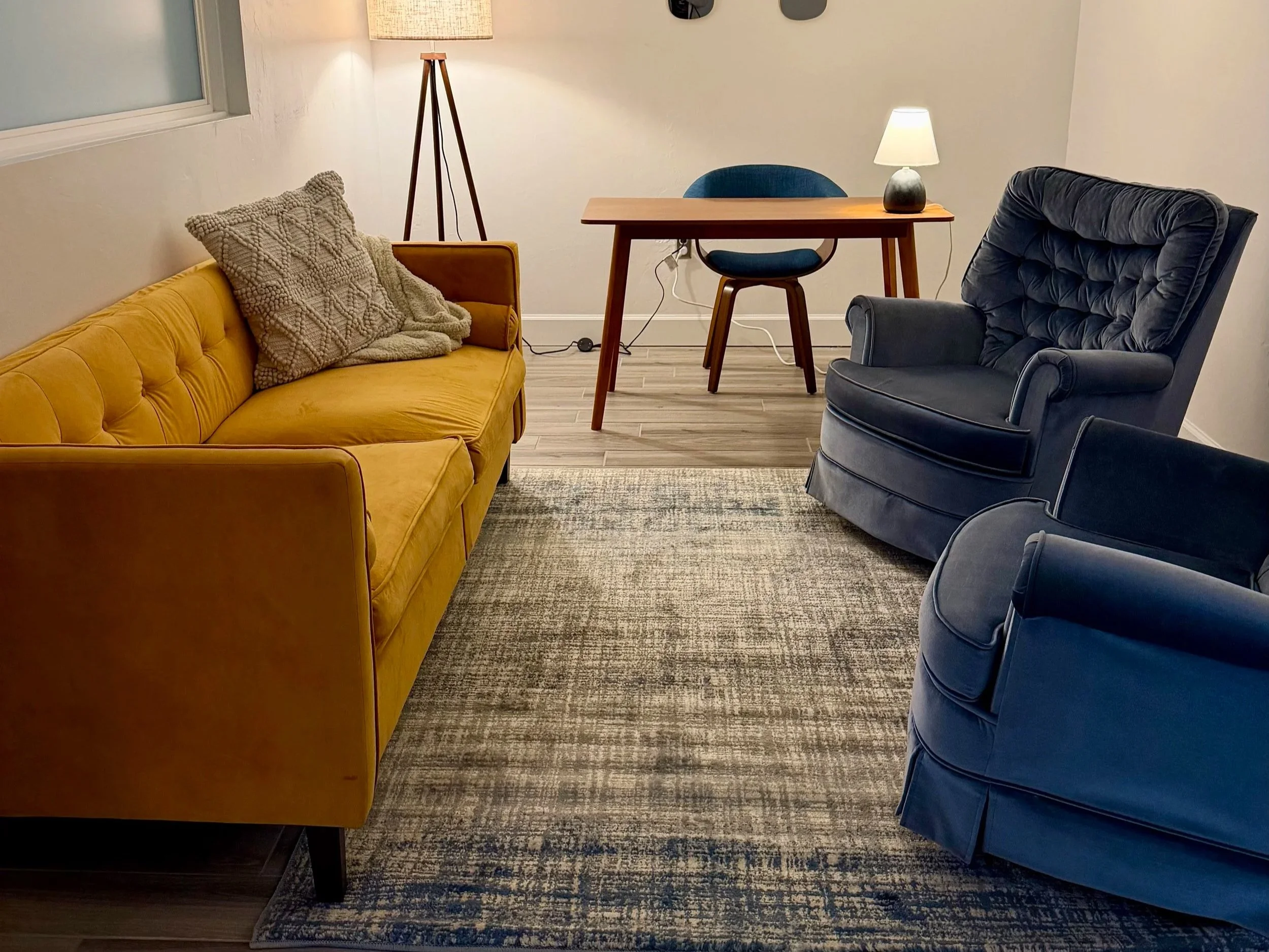 A living room with a mustard yellow sofa, a dark blue armchair, a matching dark blue chair, a mid-century wooden desk with a small lamp, a floor lamp with a tripod stand, and a textured area rug on wooden flooring.