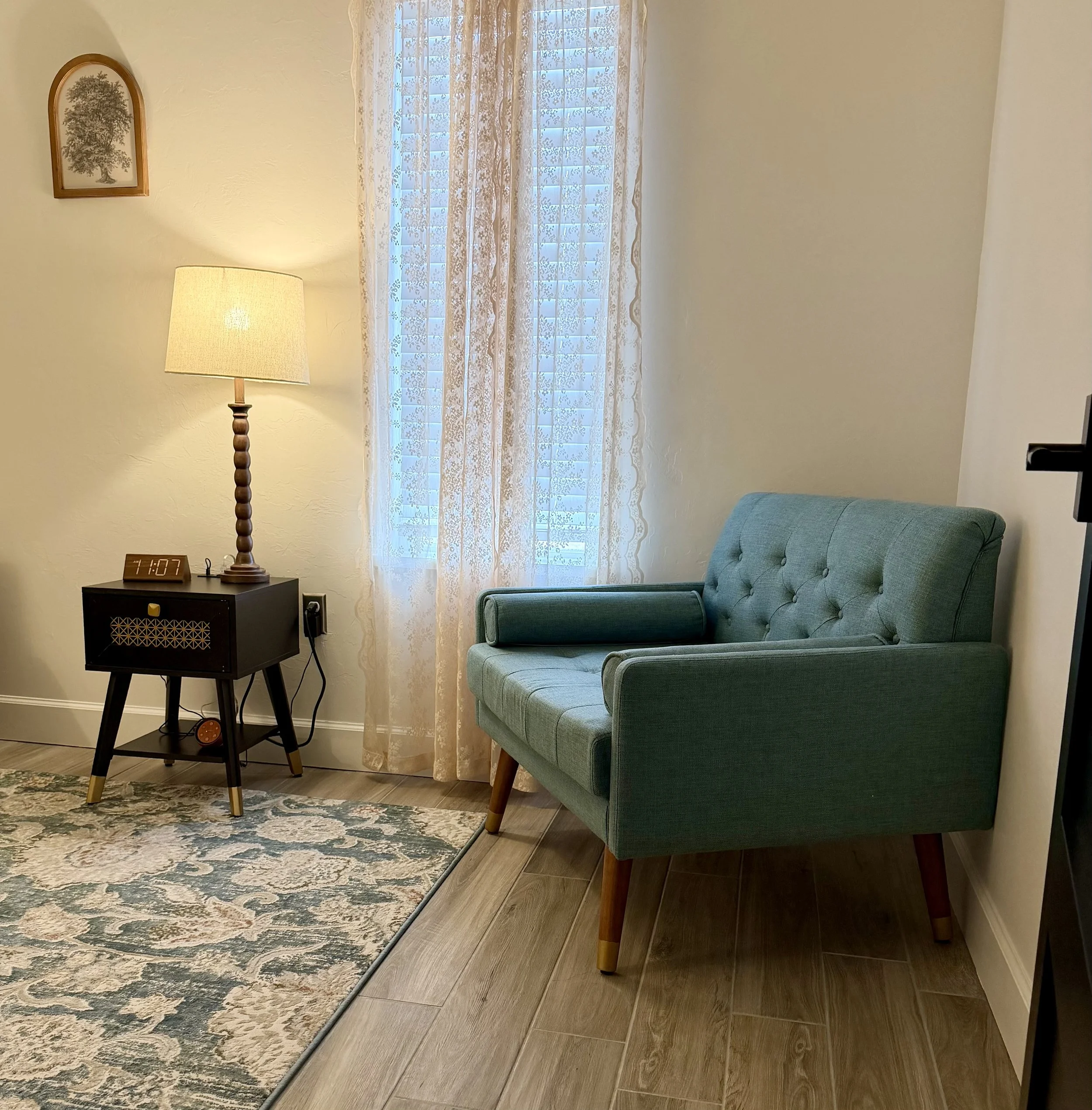 Living room corner with a green mid-century modern sofa, a side table with a digital clock and lamp, a lace curtain covering a window, and a wall picture of a tree.