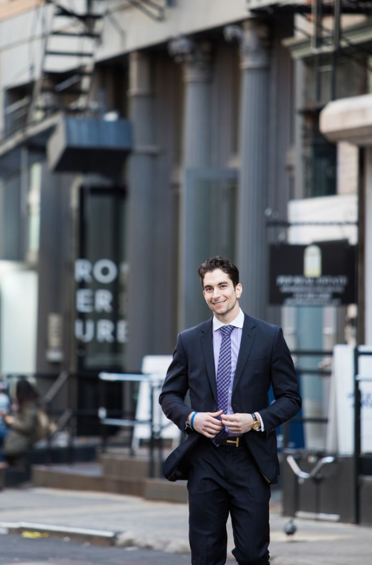 A young man in a business suit walking on an urban sidewalk, smiling, with modern buildings in the background.