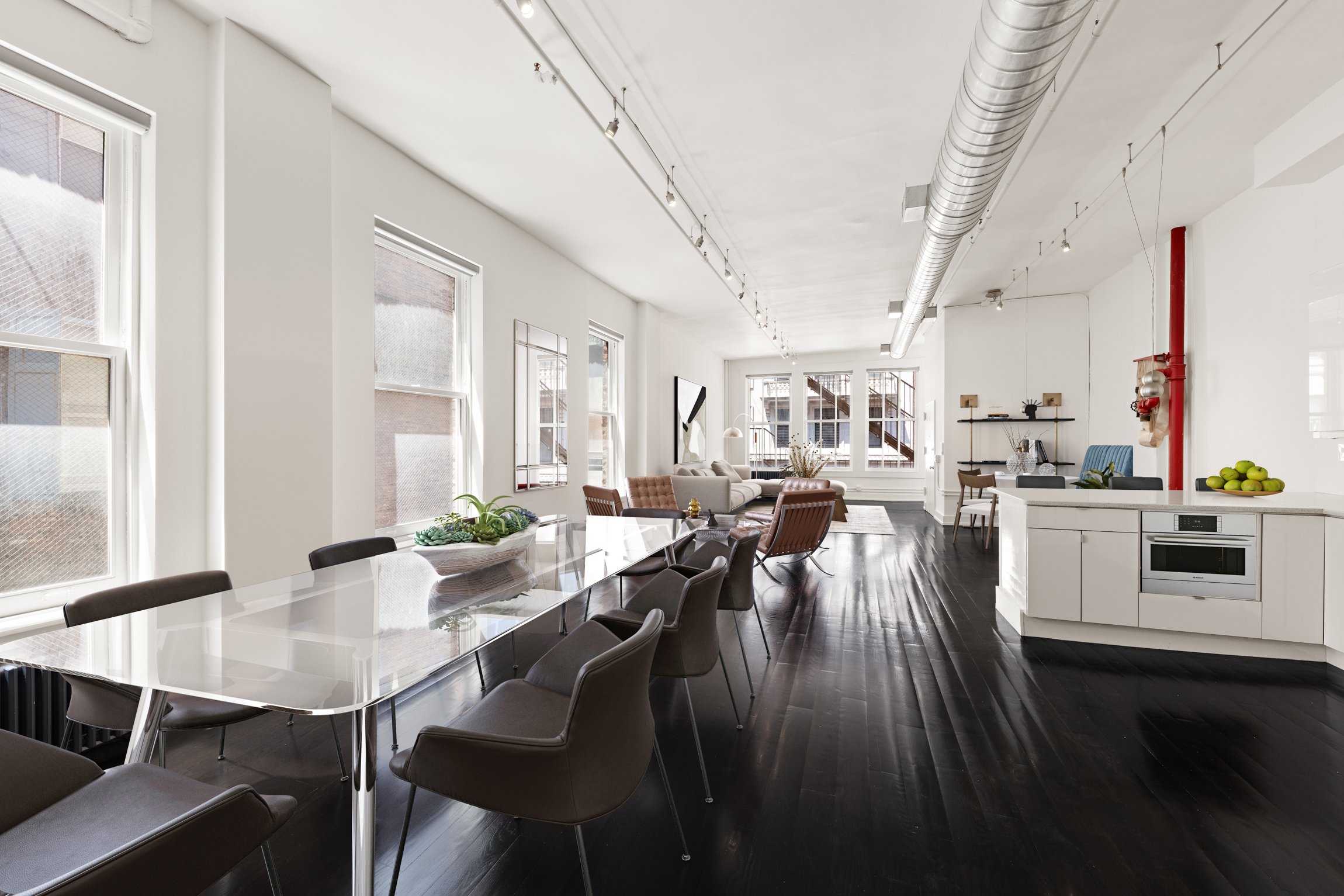 Open-concept living space with dining area, seating area, and kitchen. Large windows, dark wooden floors, white walls, modern furniture, and decorative items.