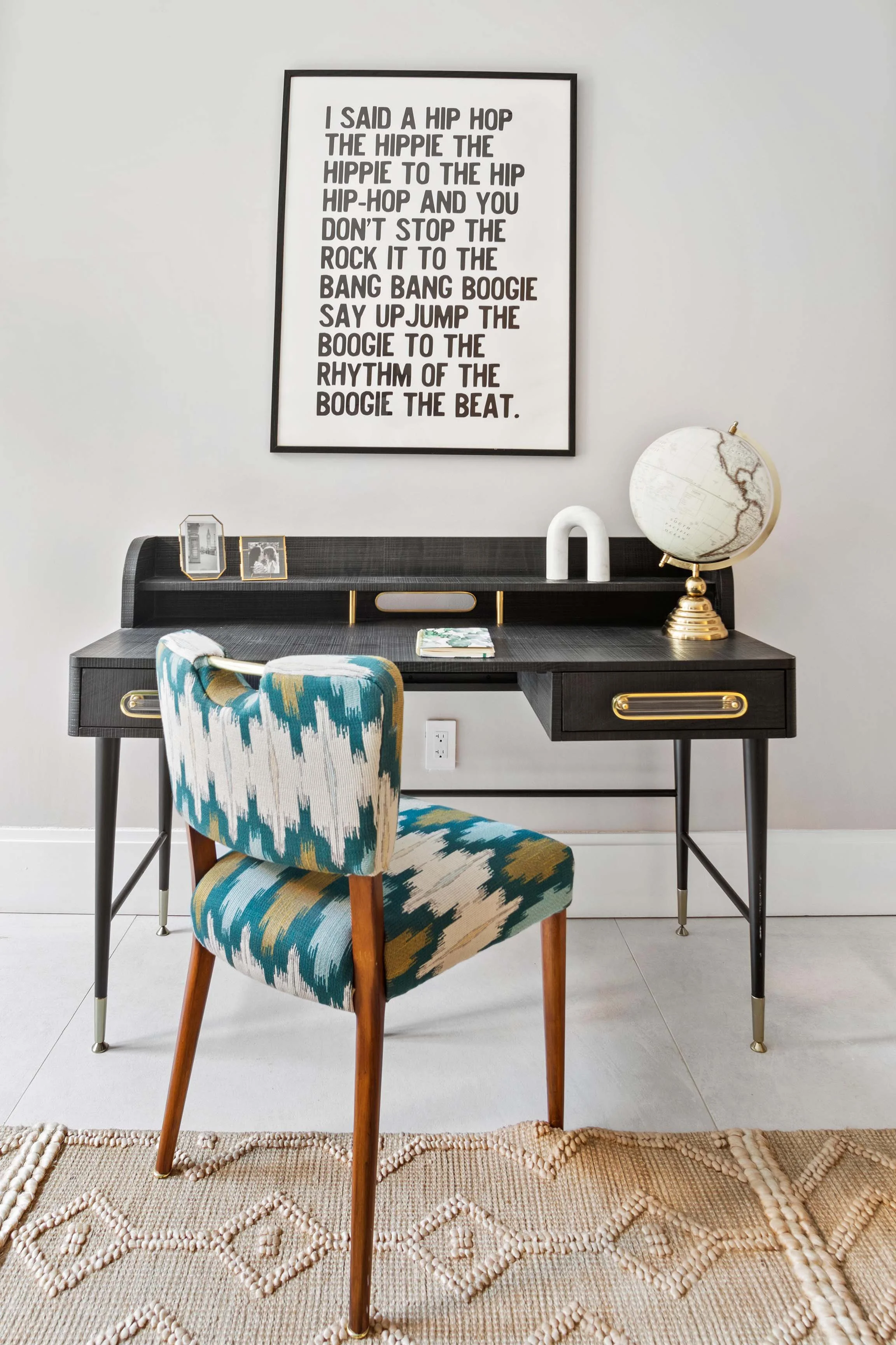 A black desk with gold accents, a globe, and picture frames, set against a white wall with a large framed typography print, and a patterned upholstered chair in front, on a beige textured rug.