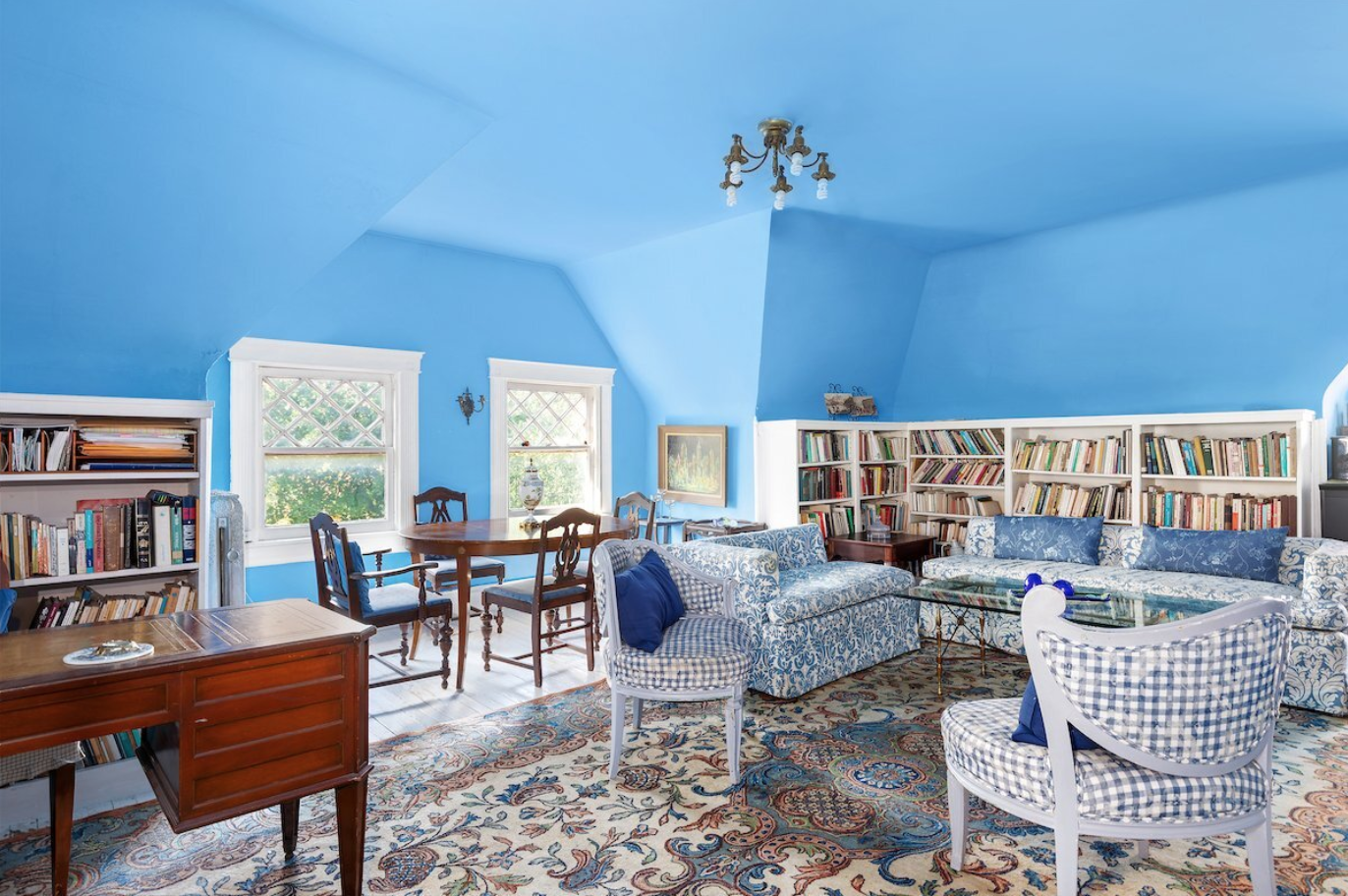 A cozy living room with blue walls, white trim, and a patterned area rug. It features a mix of vintage and classic furniture including a blue patterned sofa, checkered armchairs, a wooden dining table with chairs, and bookshelves filled with books. The room has three windows allowing natural light and a small chandelier hanging from the ceiling.
