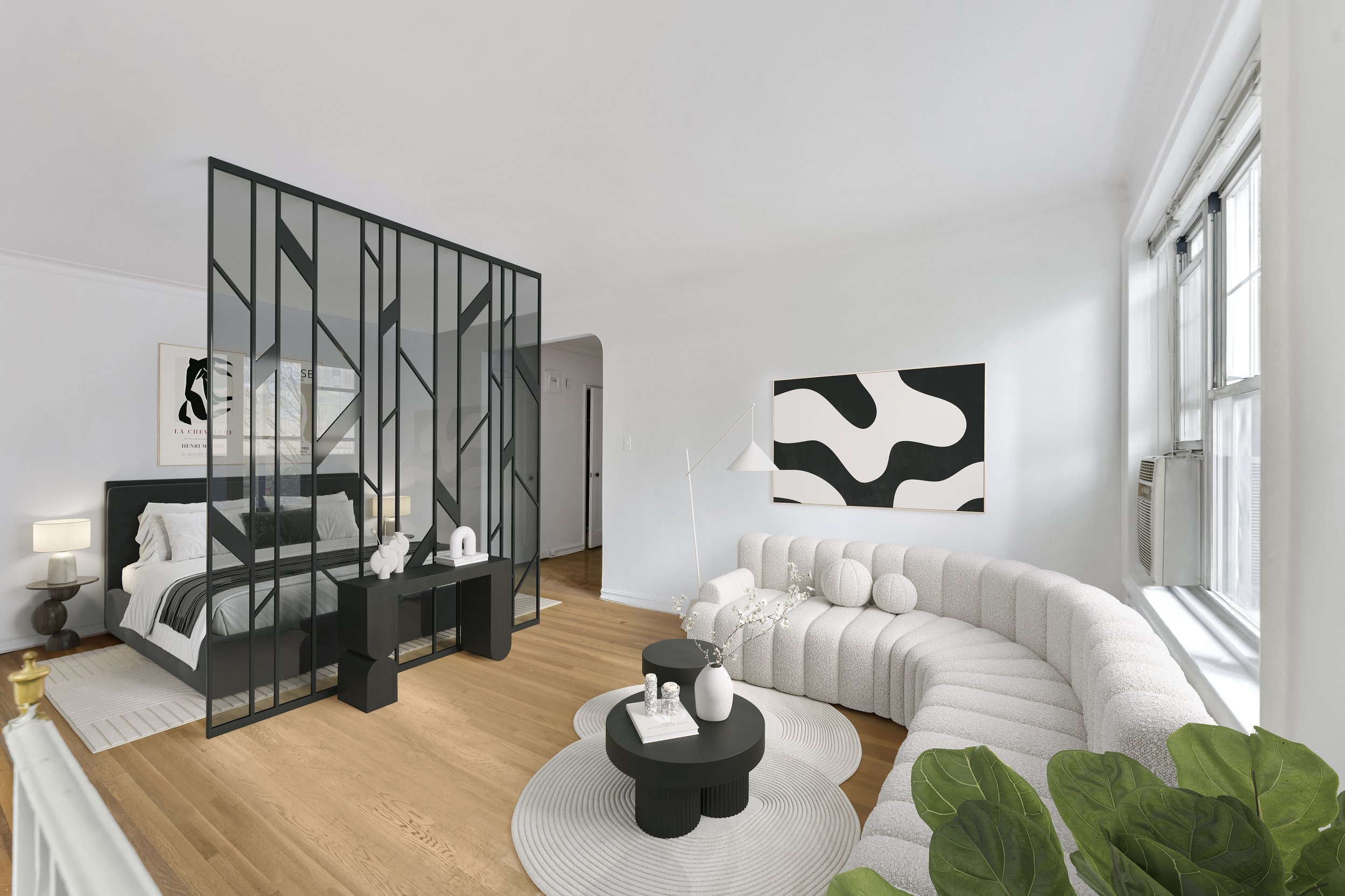 Modern living room with white curved sofa, black and white abstract wall art, black coffee table with vases, large windows, and a bedroom separated by a black metal room divider.
