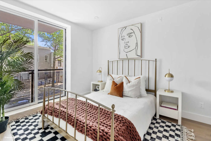 Modern bedroom with white walls, a gold metal bed frame, white bedding, and a rust-colored throw blanket. Two white nightstands with gold lamps flank the bed. A large abstract line art portrait hangs above the bed. A sliding glass door with a view of a backyard and neighboring houses is to the left. A black and white checkered rug covers part of the light wood floor, with a tall green plant in a black pot near the window.