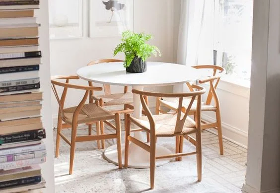 A white round dining table with a green plant in a black pot center and six wooden chairs in a bright room with large windows.