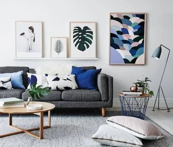 Living room with a gray sofa, blue and patterned cushions, a wooden coffee table, and a side table with potted plants. Framed artwork of leaves and an abstract ocean wave on the wall, and a floor lamp.
