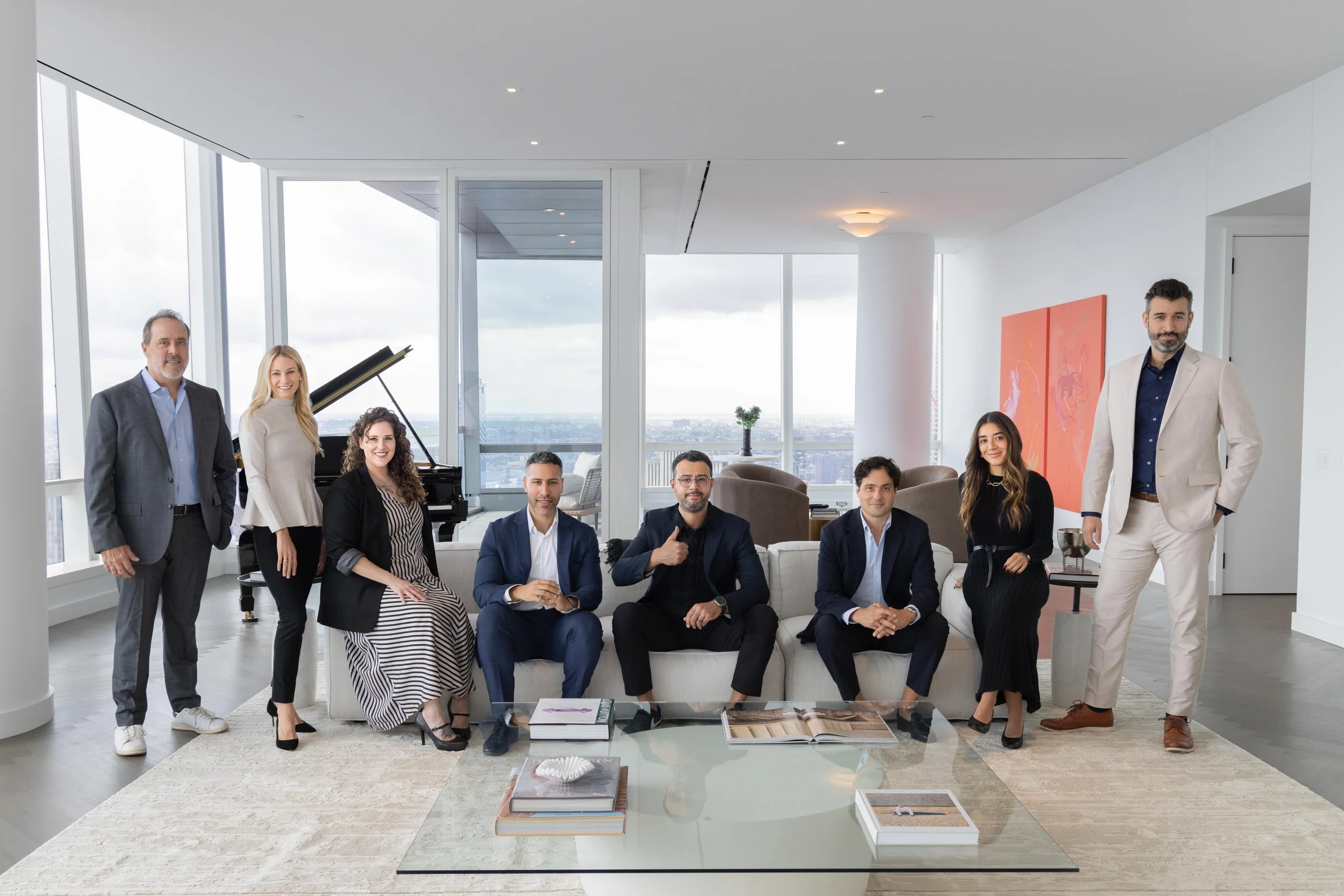 A group of nine professionally dressed people in a modern high-rise apartment with large windows and a city view, sitting and standing around a coffee table with books and decorative items.