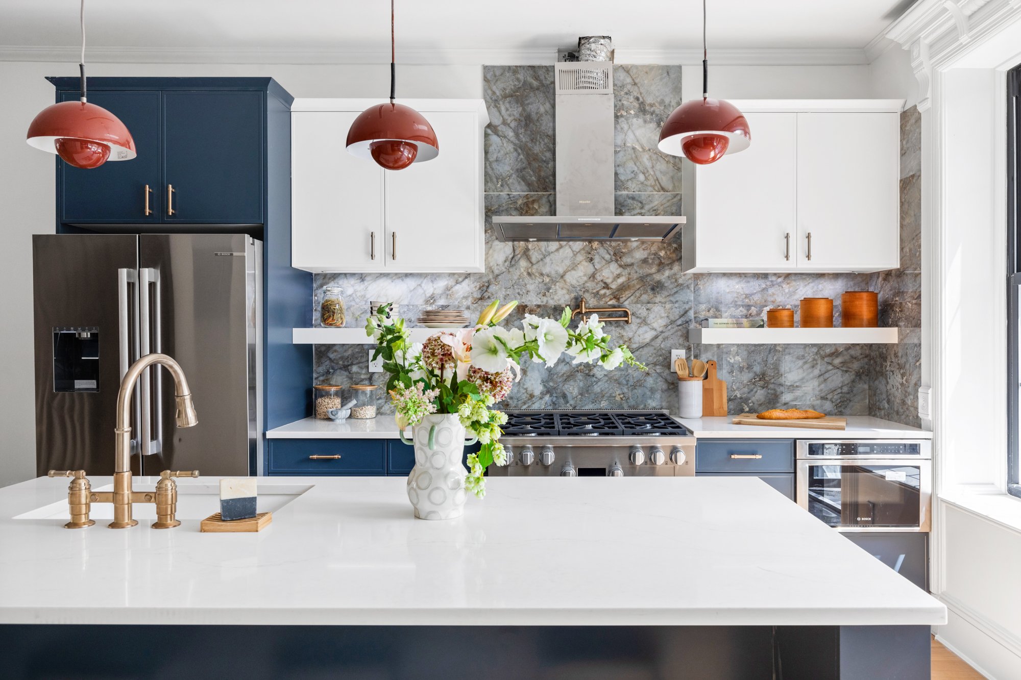 Modern kitchen with white countertops, blue and white cabinets, stainless steel refrigerator, gas stove, and a vase of flowers on the island. Red pendant lights hang above the kitchen island, and a window brings natural light.