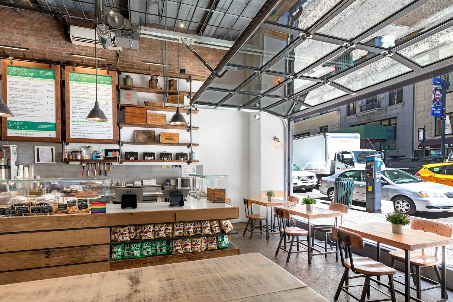 Interior of a modern cafe with wooden tables, chairs, and potted plants near large open windows showing parked cars and city street outside.