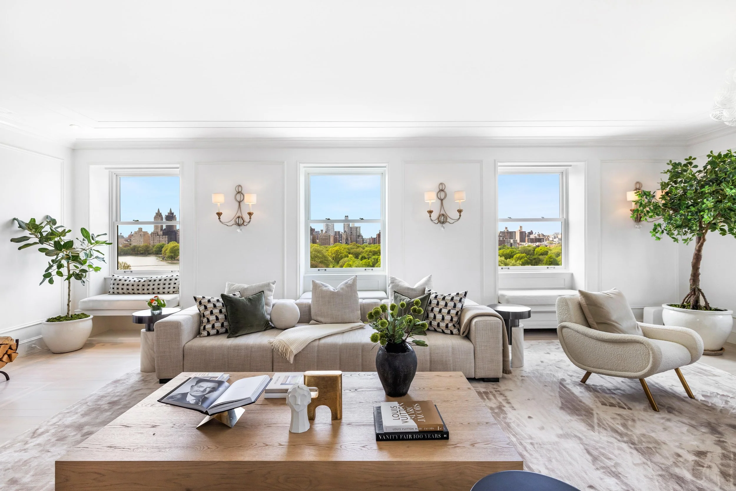 Bright living room with white walls, three large windows showing city skyline and green trees, elegant beige sofa with assorted pillows, modern armchair, wooden coffee table with decor and books, large potted plants, and decorative wall sconces.