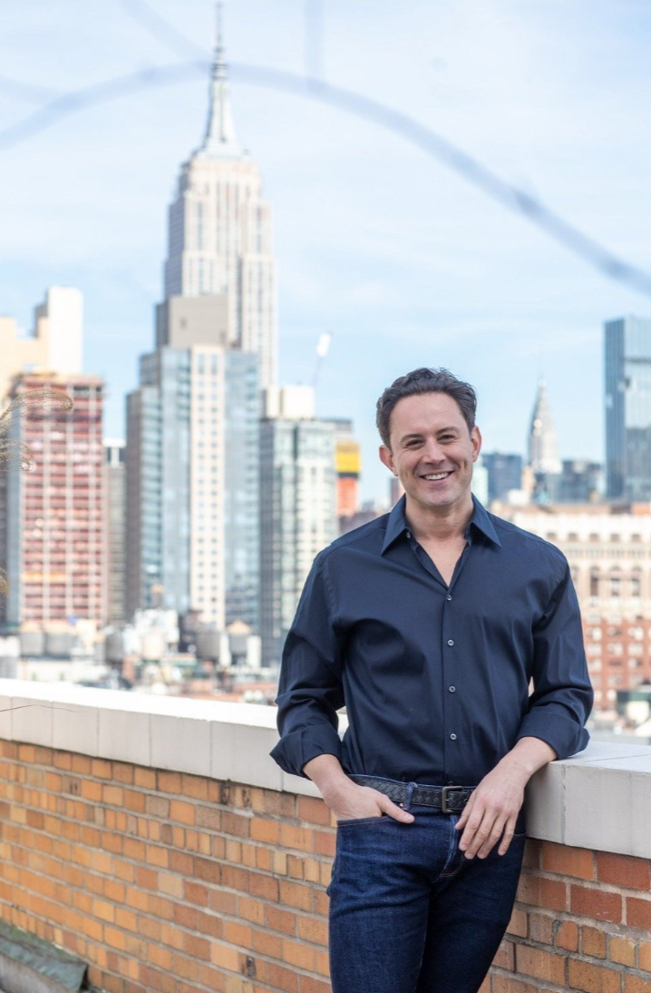 A man smiling on a rooftop with the New York City skyline, including the Empire State Building, in the background.