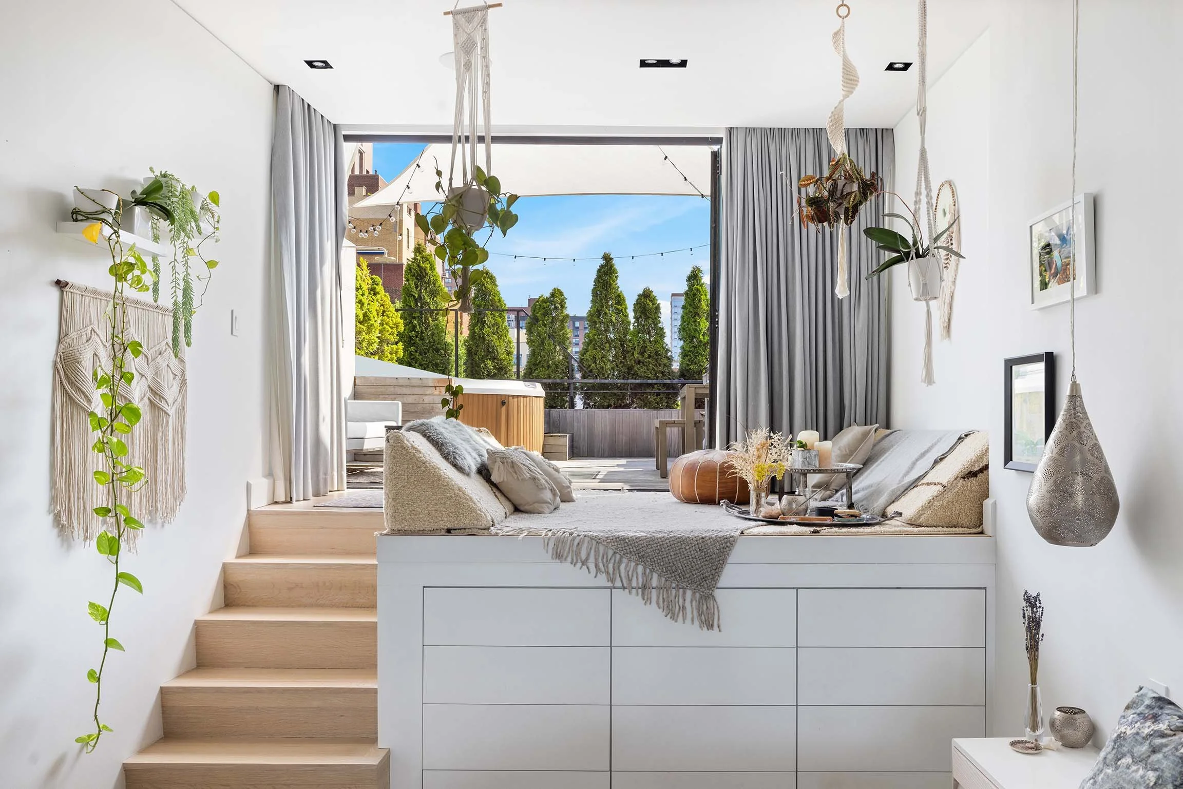 Bright sunroom with a bay window opening to a balcony with outdoor seating, decorated with hanging plants, pampas grass, candles, and cozy cushions, overlooking lush greenery and neighboring buildings.