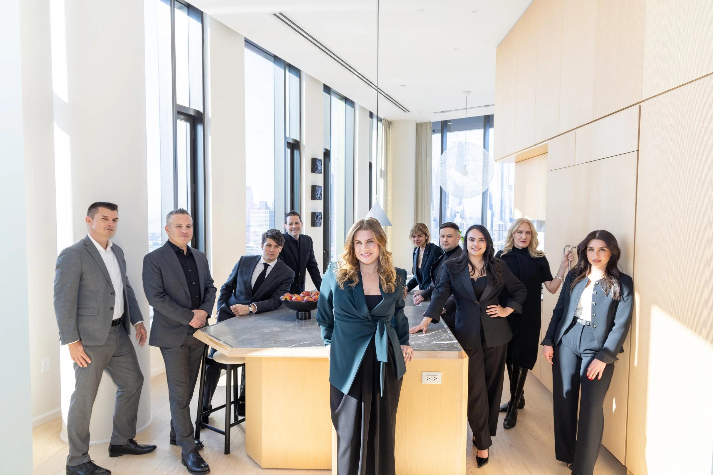 A group of nine business professionals, six women and three men, standing in a modern office kitchen with large windows, a marble table, and cityscape views. They are smiling and dressed in formal business attire.
