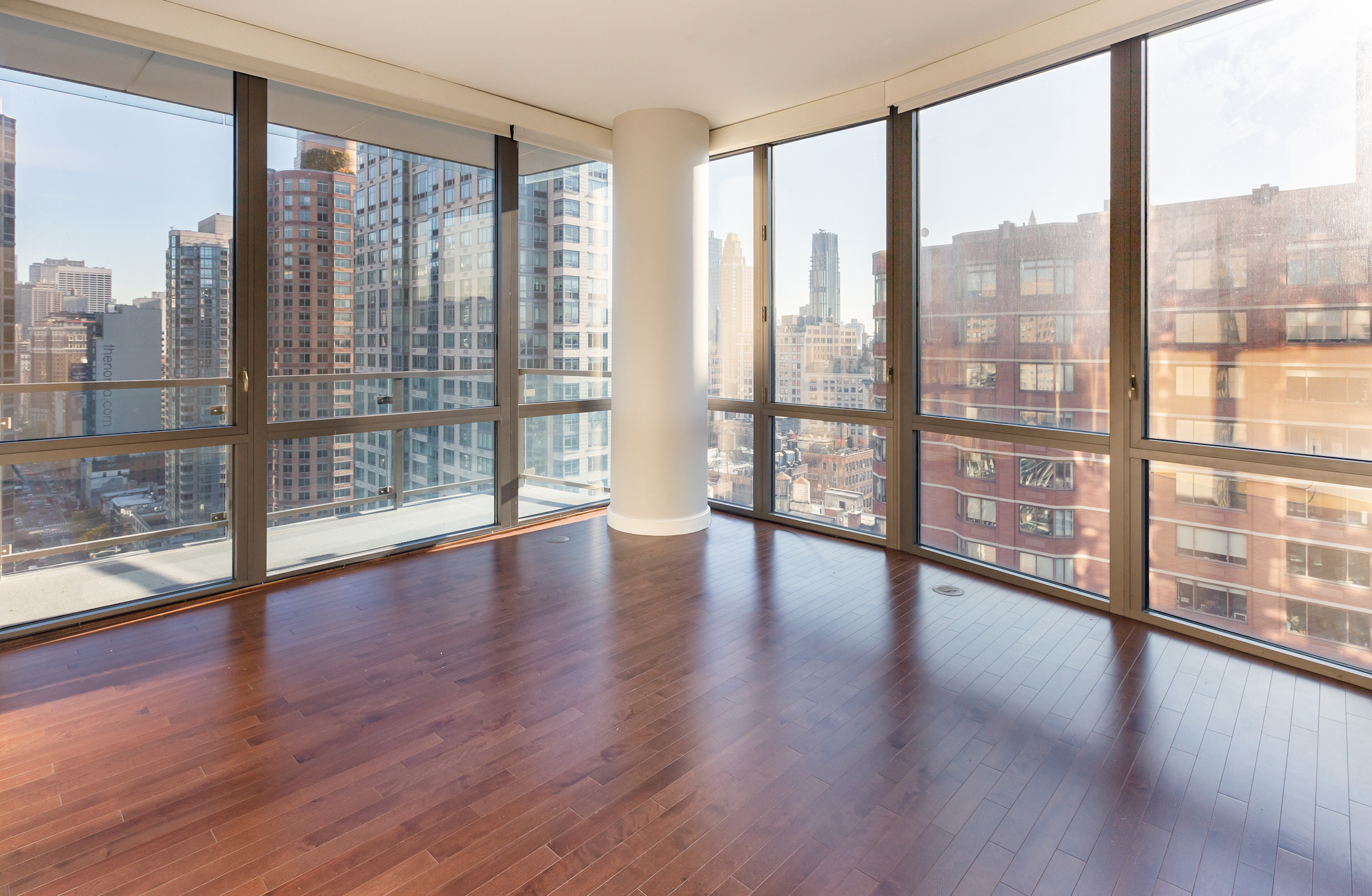 Empty apartment room with large floor-to-ceiling windows overlooking a city skyline with tall buildings, and a wooden floor and a white column in the corner.