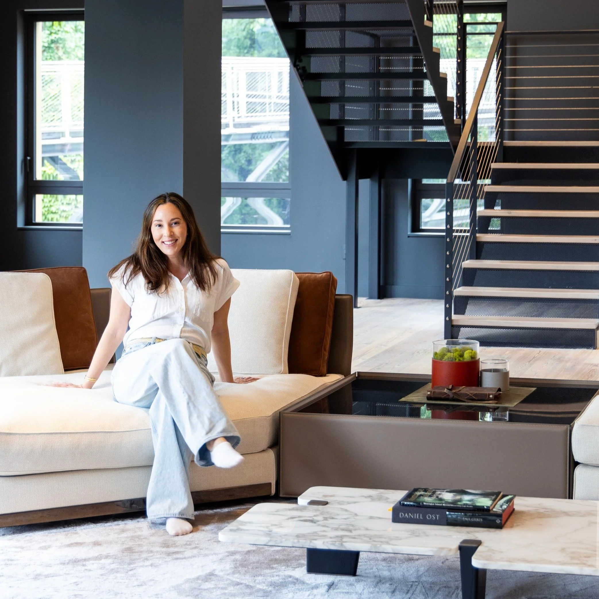 A woman with long dark hair wearing a white shirt and light-colored pants sitting on a beige couch in a modern living room with large windows, a staircase with metal railings in the background, and a marble coffee table with books on it.