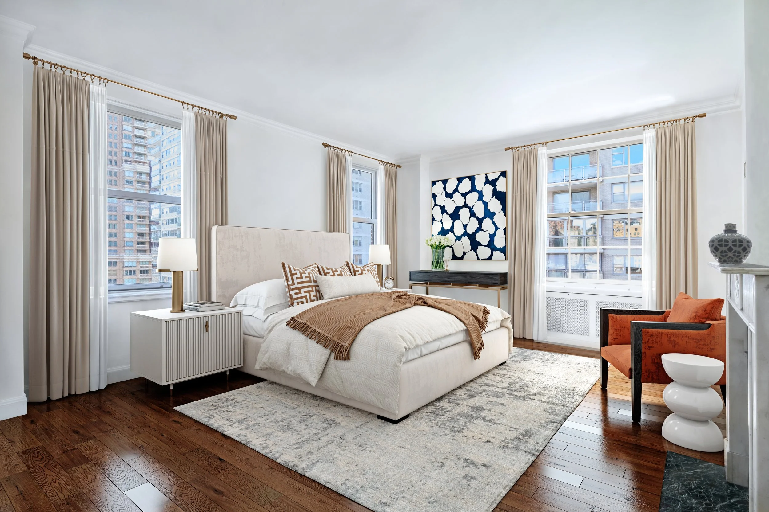 Bright bedroom with large windows, beige curtains, white walls, wooden floor, and modern furniture. There is a bed with white and beige bedding, patterned pillows, a nightstand with a lamp, a chair with orange upholstery, and wall art featuring white