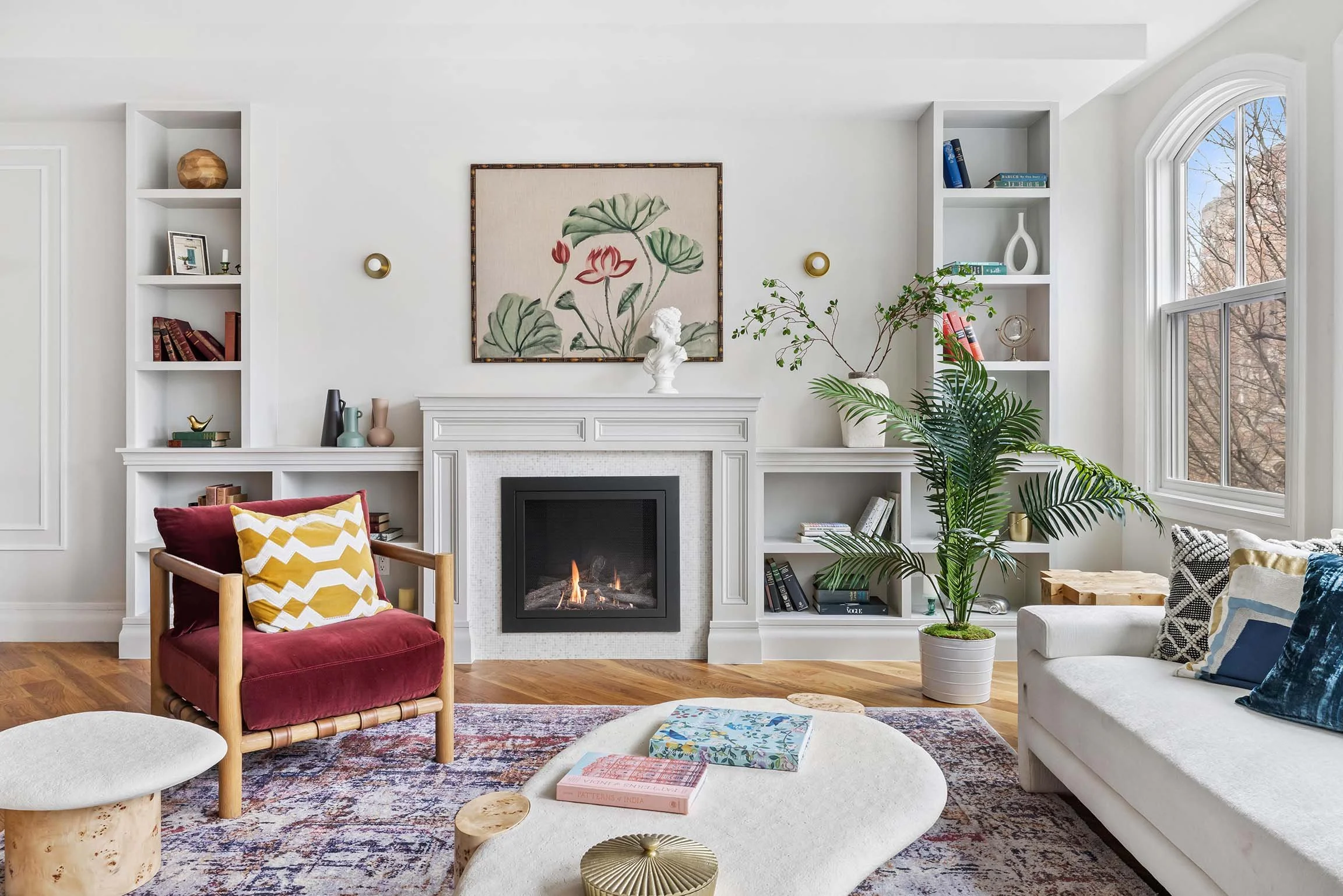 Living room with white cabinetry, a fireplace, large window, and modern decor including plants, artwork, and cozy furniture.