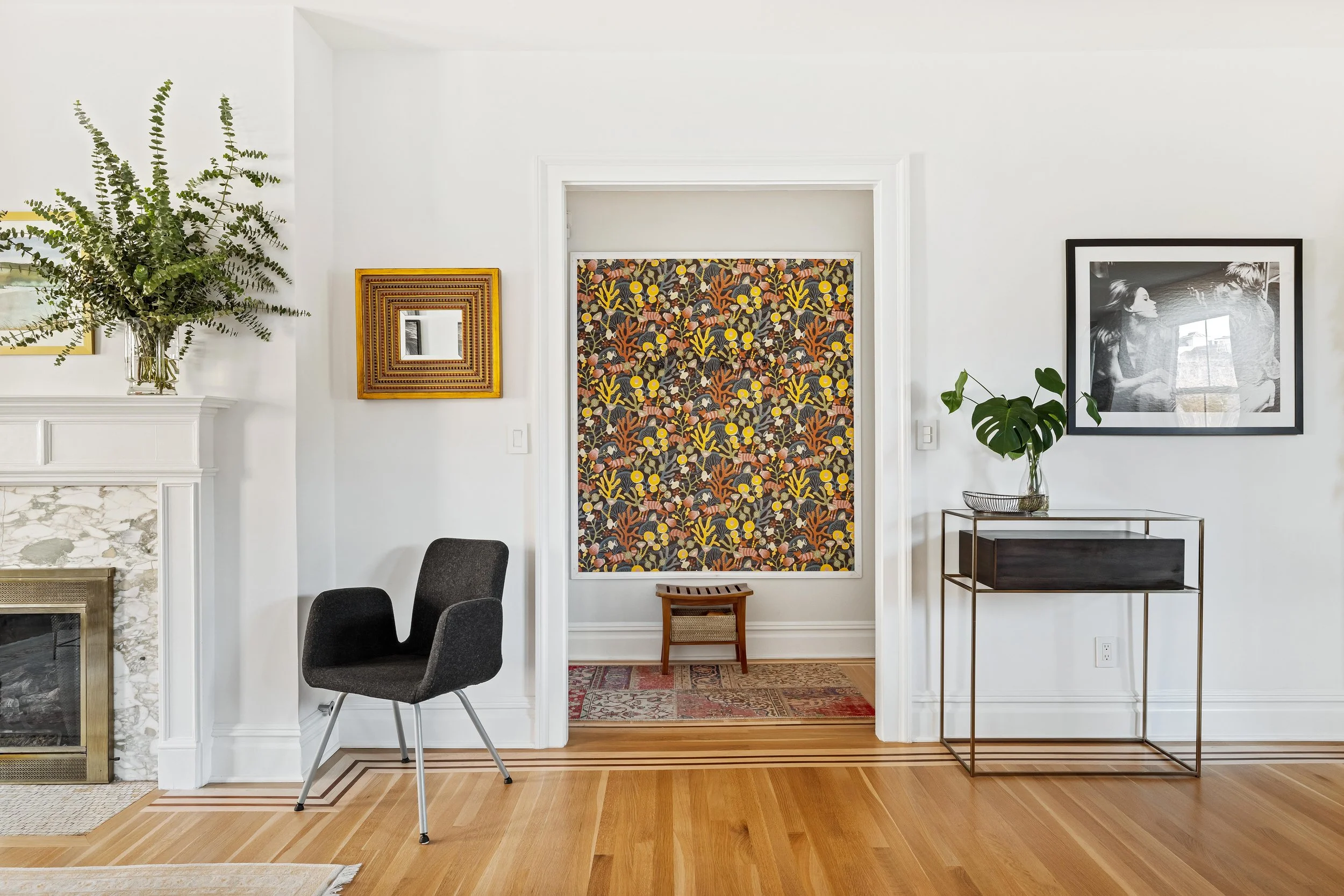 Living room with white walls, a black chair, a fireplace with a marble mantle, a large plant in a vase, framed art, and a doorway with colorful patterned wallpaper.