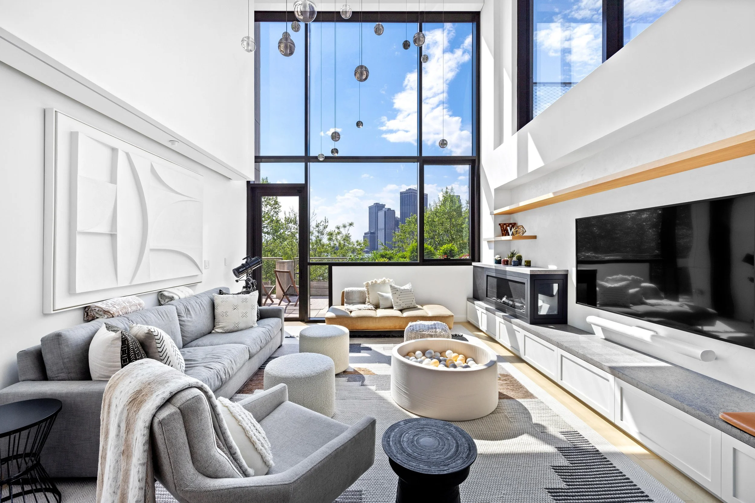Modern living room with large windows, gray sofas, white decor, and city skyline view.