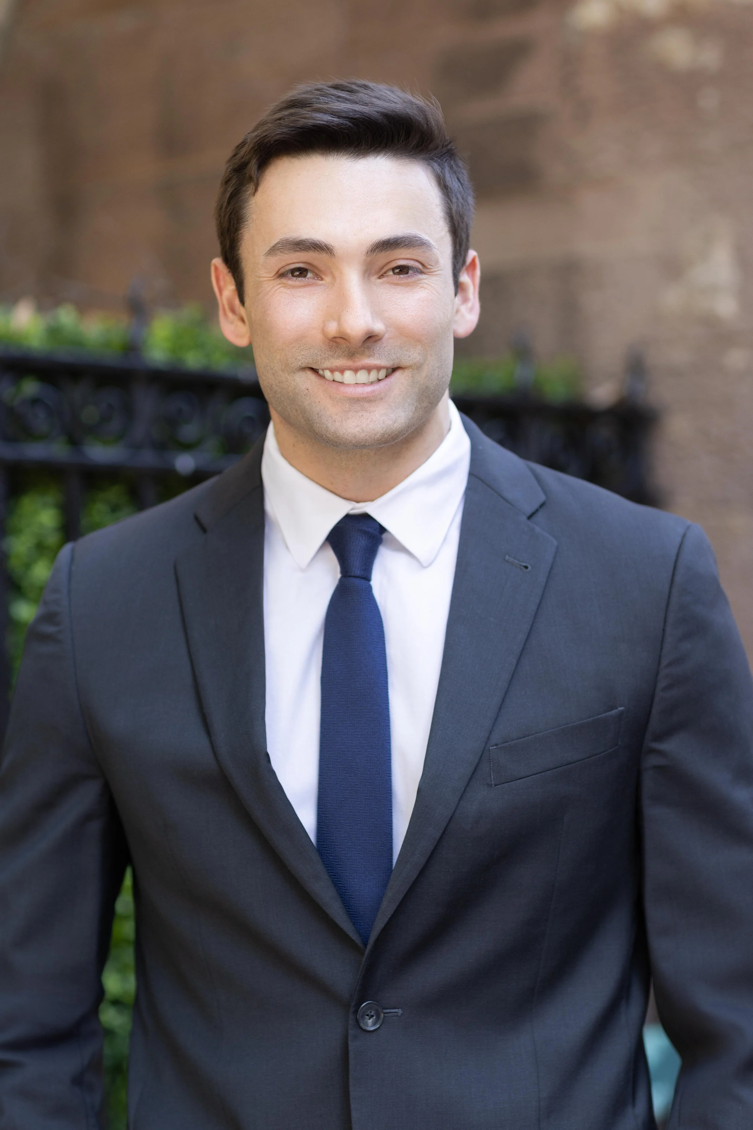A young man wearing a black suit, white shirt, and dark blue tie, smiling outdoors in front of a background with a brick wall and greenery.