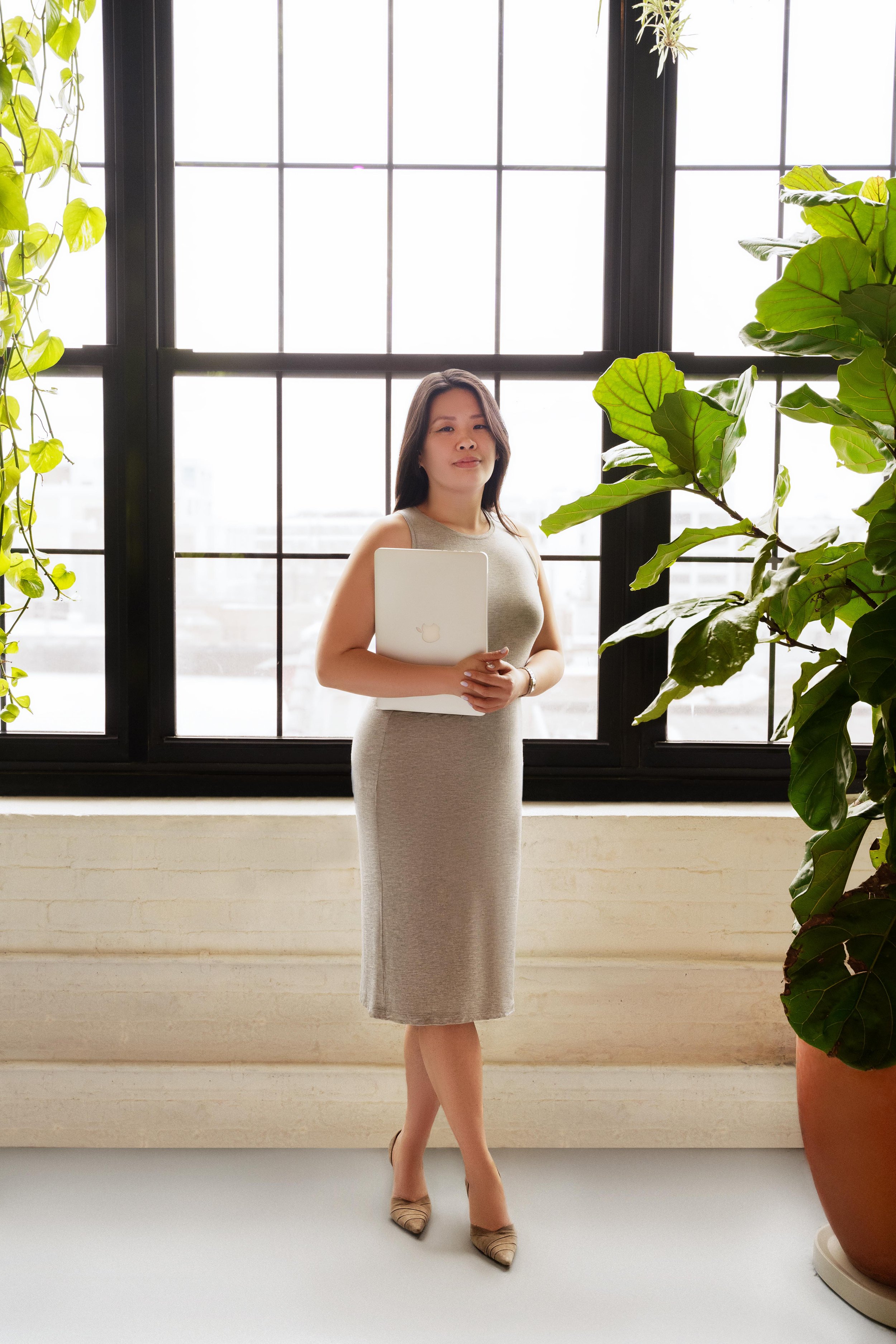 A woman in a beige dress standing in front of large window with plants on either side, holding a closed laptop.