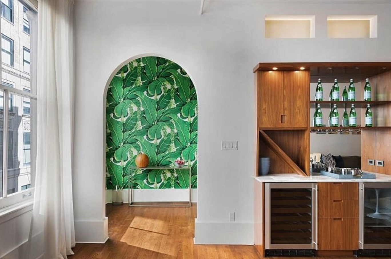 Interior view of a modern room with a bác̣klit bar area featuring wood cabinets, glass bottles, and a wine cooler. There is a large window with white curtains and an arched nook with green banana leaf wallpaper, a small table with a decorative vase and flower, and hardwood flooring.