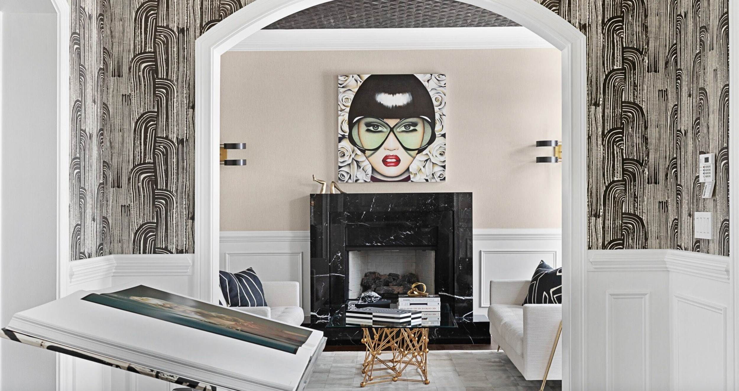 Living room with a fireplace, modern art portrait of a woman with glasses above the mantel, black marble fireplace, black and white striped books, gold decorative sculpture, two white armchairs with black and white pillows, and patterned wallpaper