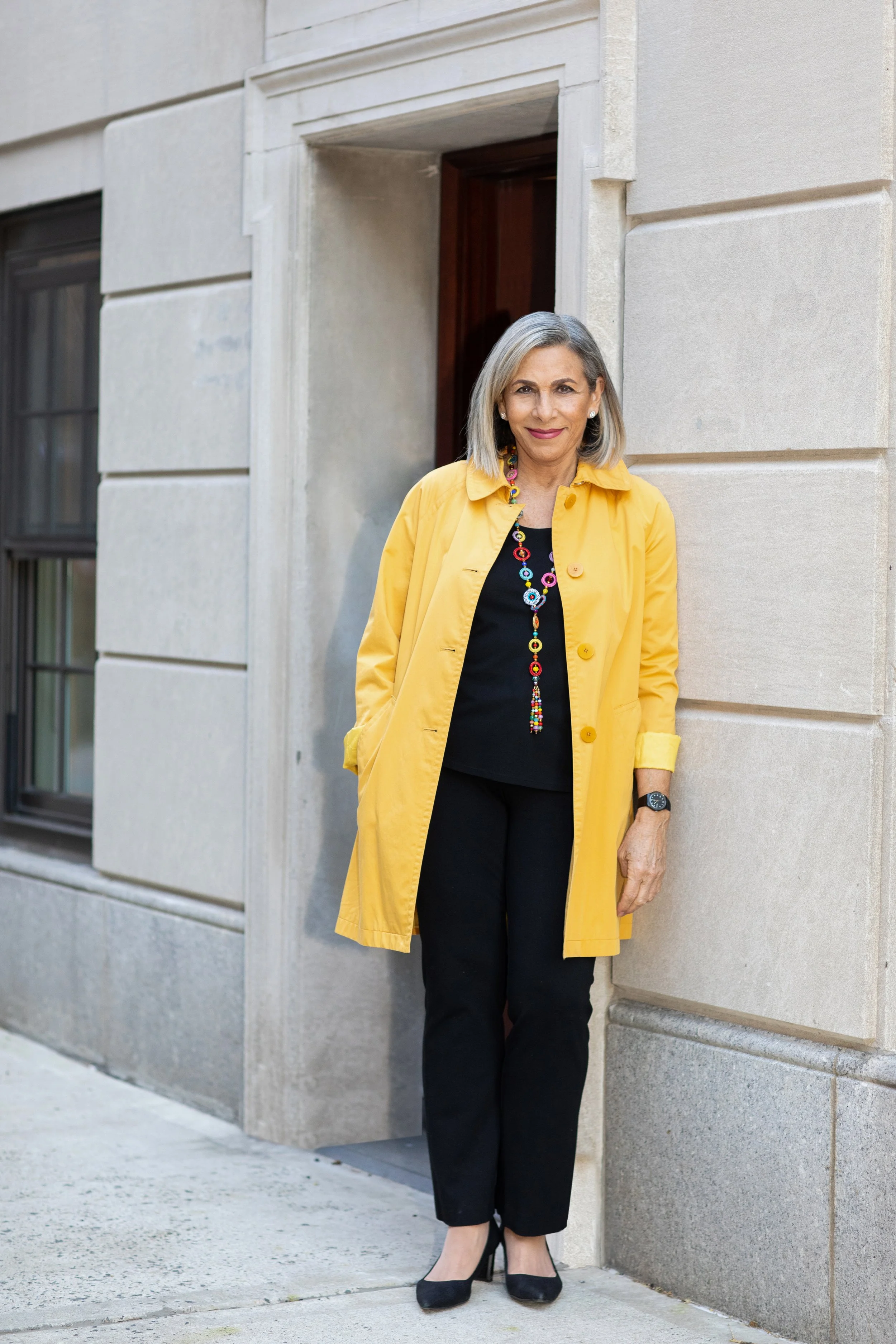An older woman with gray hair wearing a yellow coat, black top, and black pants, standing outside a building with stone walls.