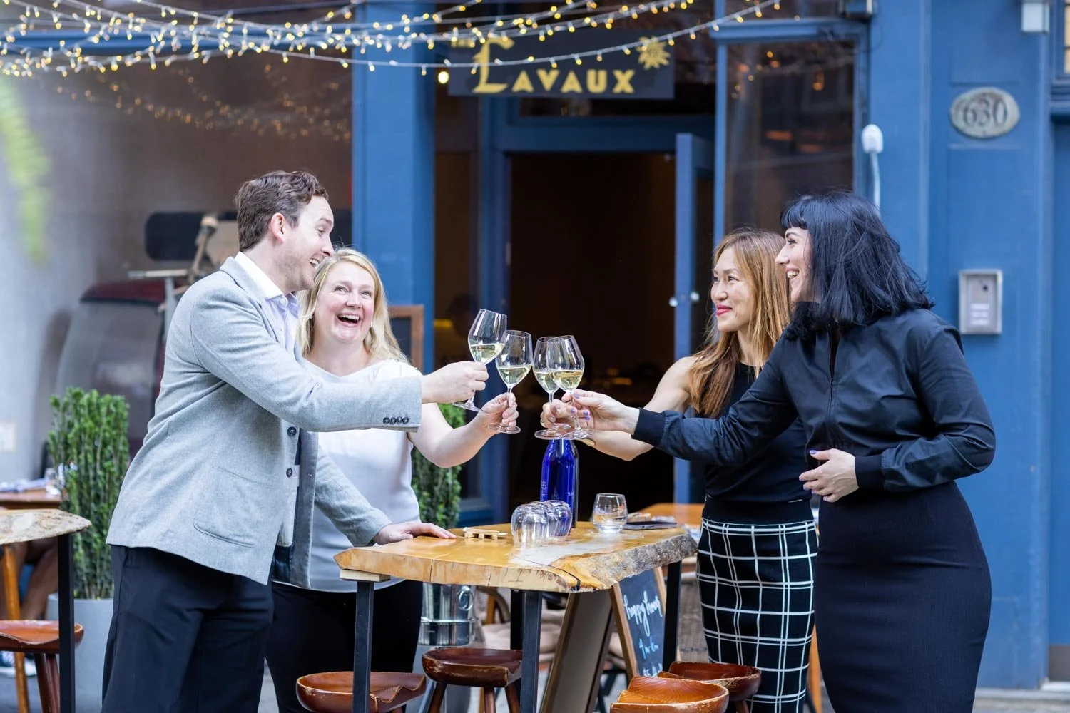 Group of four friends celebrating with wine glasses outside a restaurant with string lights and blue walls.