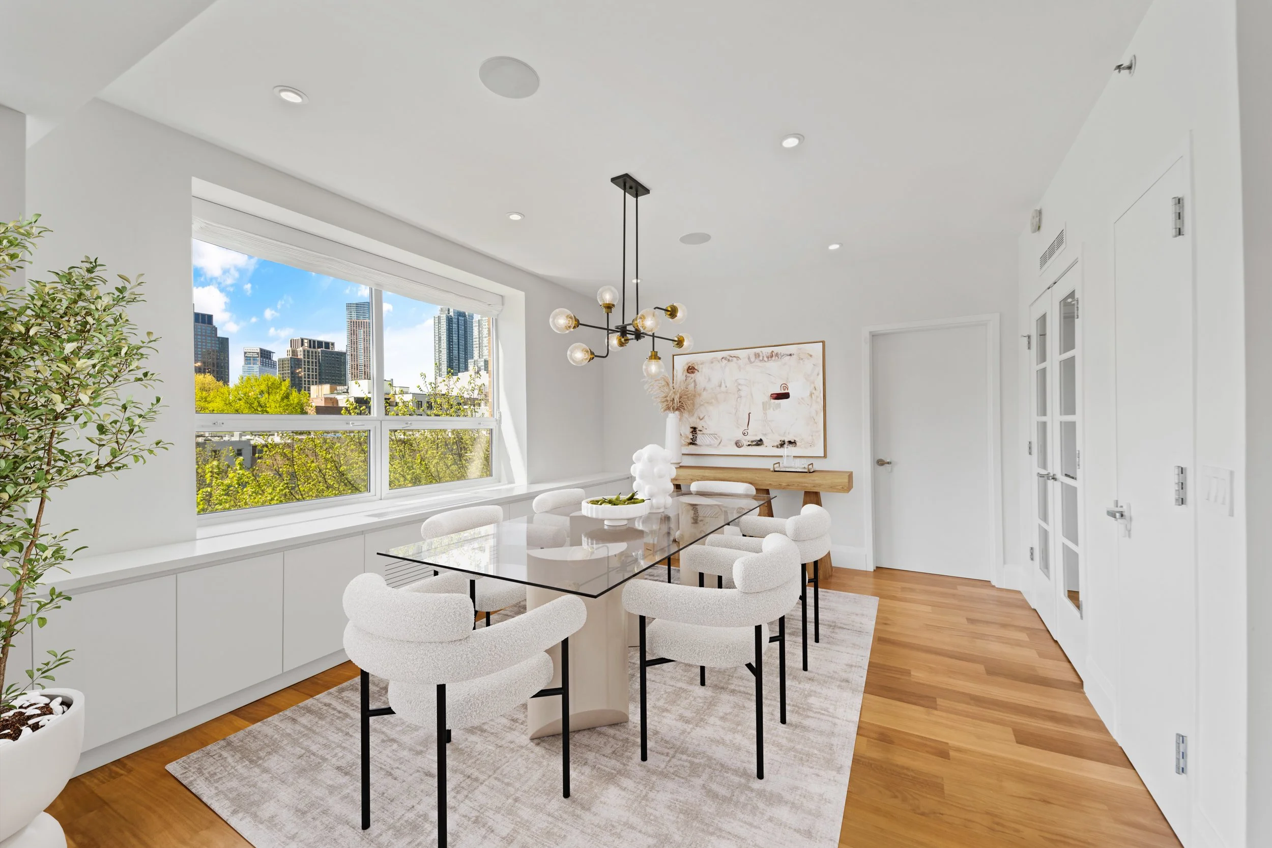 Modern dining room with a large window overlooking a cityscape, featuring a glass dining table with eight white upholstered chairs, a black and gold chandelier, and a minimalist sideboard with abstract art.