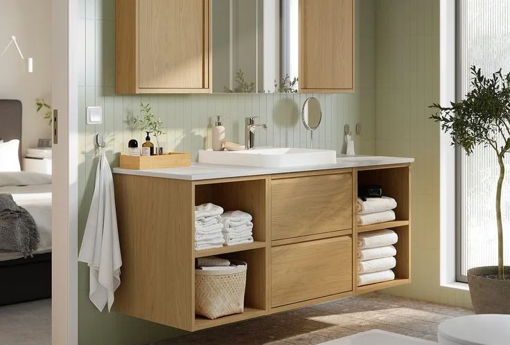 Modern bathroom vanity with a white sink, mirror, and wooden cabinets, with folded towels and bathroom essentials on the countertop, next to a window and a potted plant.