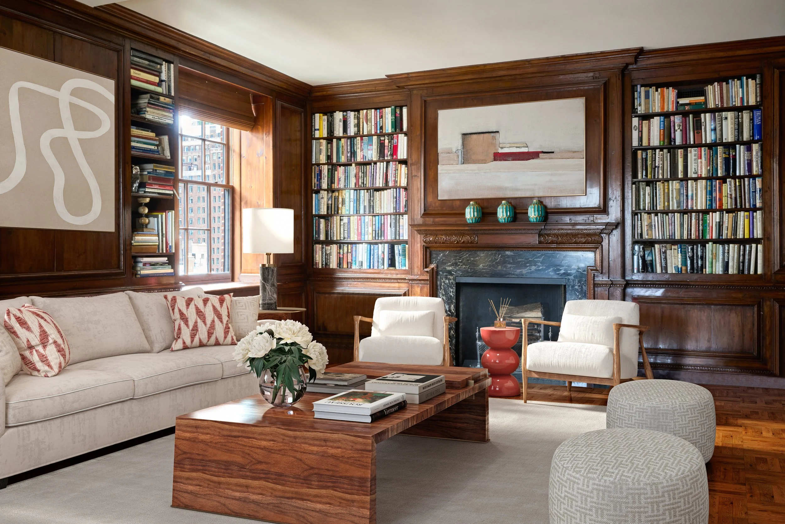 Living room with cream sofa, white armchairs, wooden walls, bookshelves, a fireplace, a wooden coffee table with books, a flower vase, and colorful decorative objects