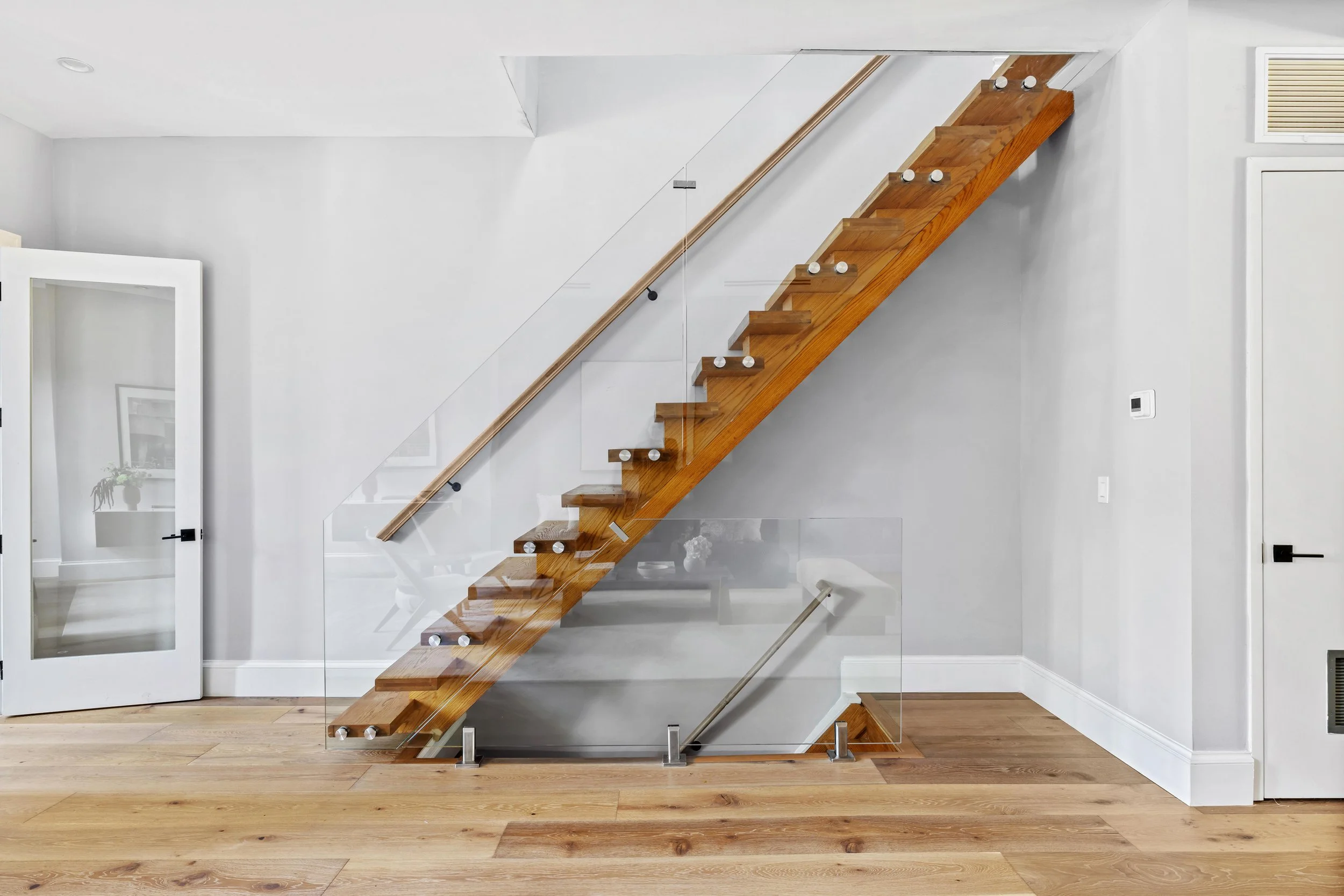 Interior of a modern home featuring a wooden staircase with glass railing, white walls, and hardwood floors.
