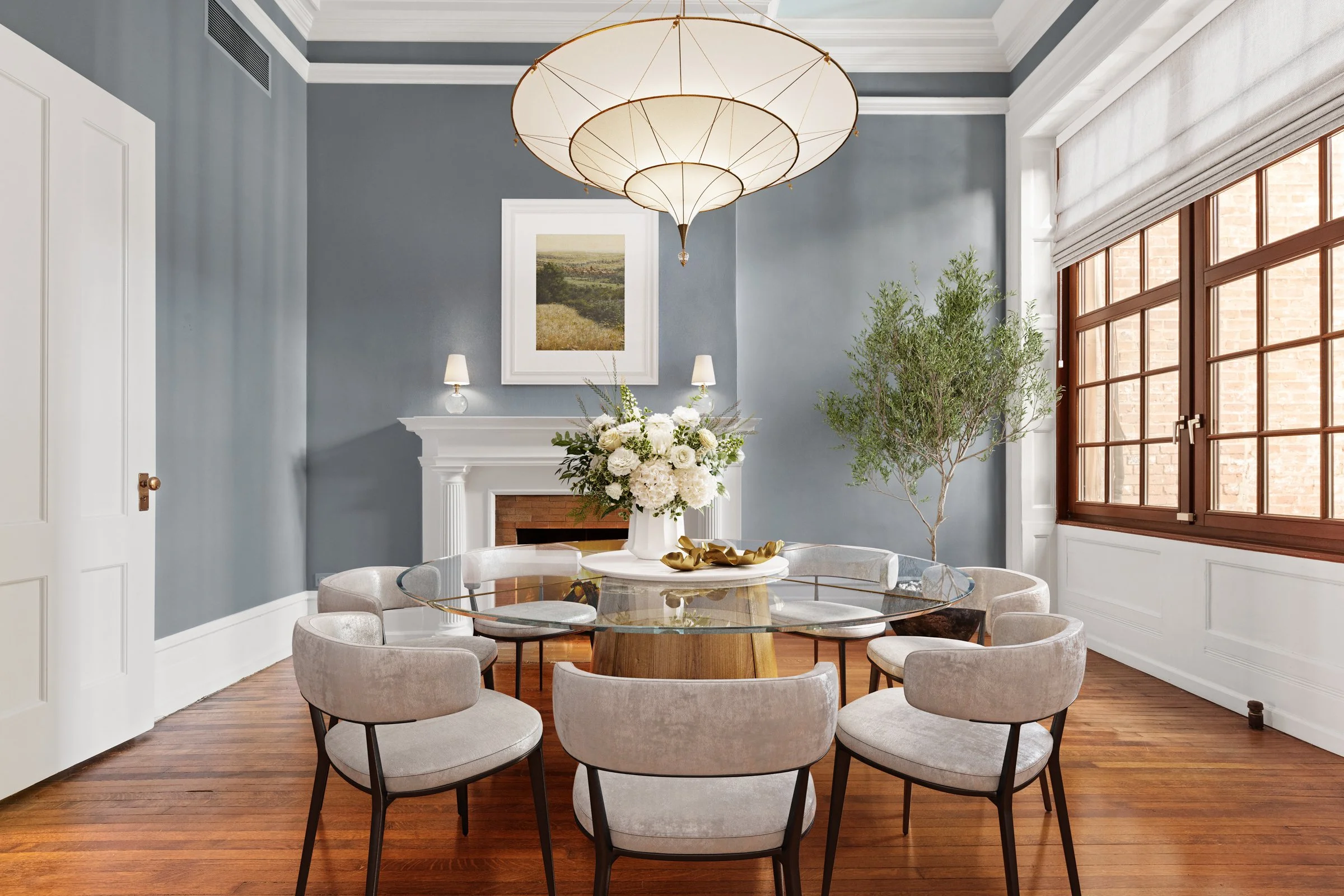 Elegant dining room with a round glass table, cream upholstered chairs, a floral centerpiece, a white fireplace, large windows with wooden frames, and a contemporary chandelier, in a room with blue walls and hardwood floors.
