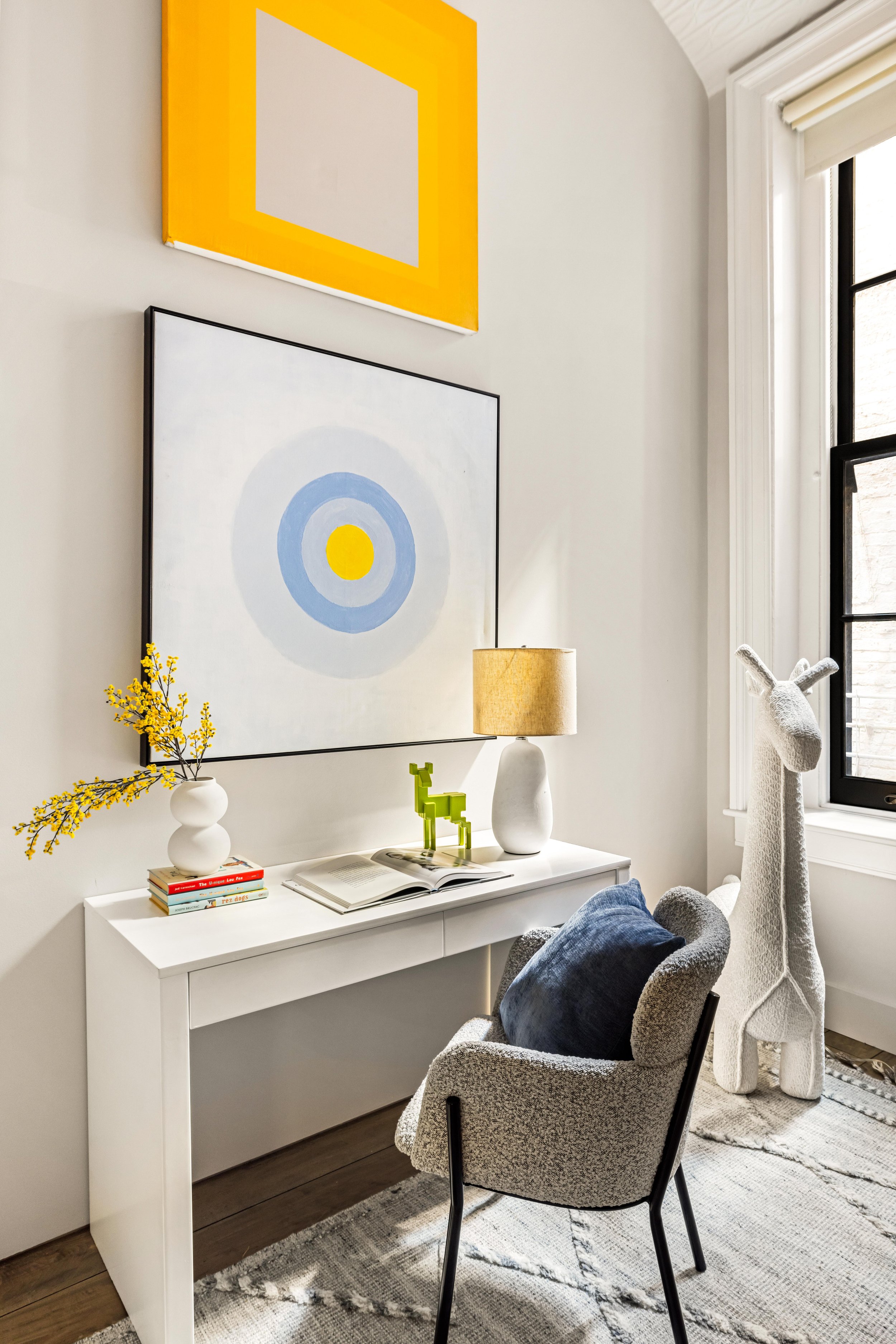 A cozy home office corner with a white desk, a gray modern chair with a dark pillow, two abstract paintings on the wall, a small white vase with yellow flowers, a stack of books, a green decorative dog figure, a table lamp, and a large stuffed giraff