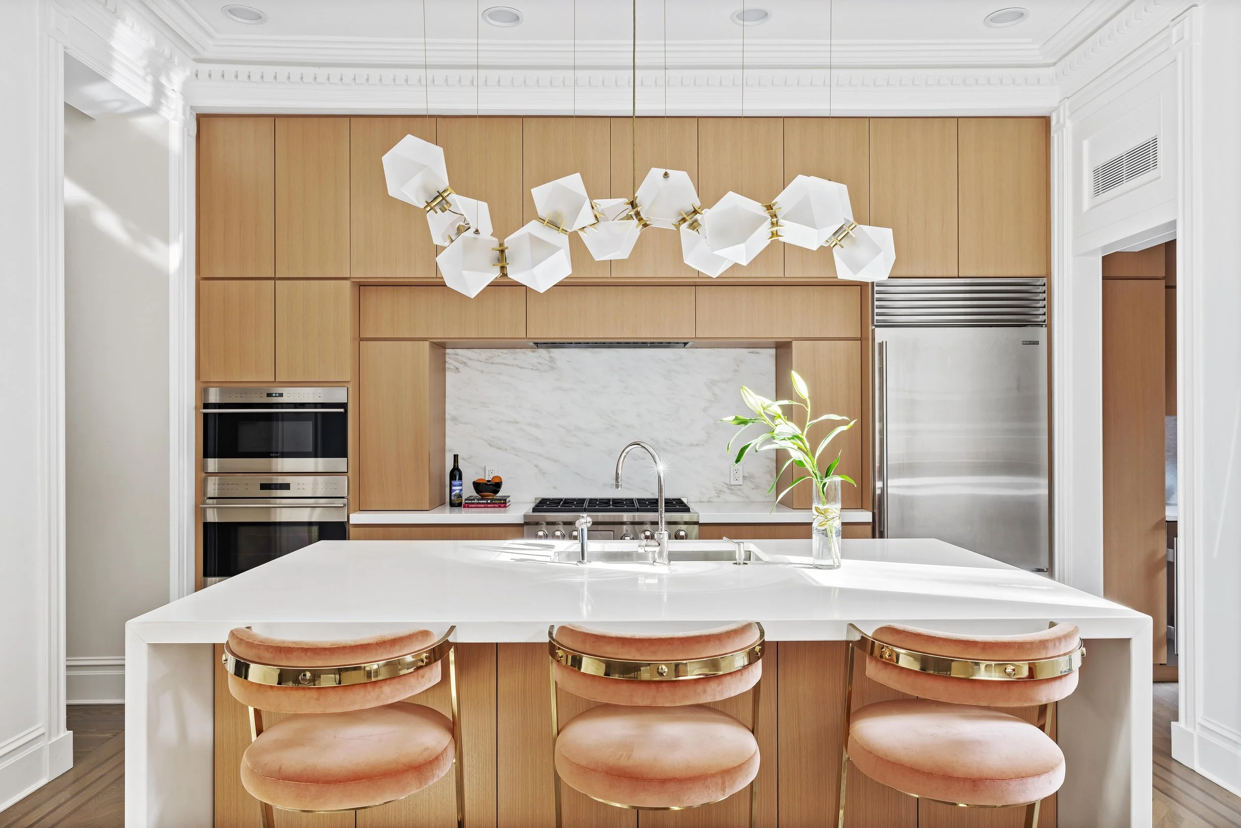 Modern kitchen with a white countertop island, light wood cabinetry, white marble backsplash, stainless steel appliances, a potted plant, and a unique white and gold chandelier.