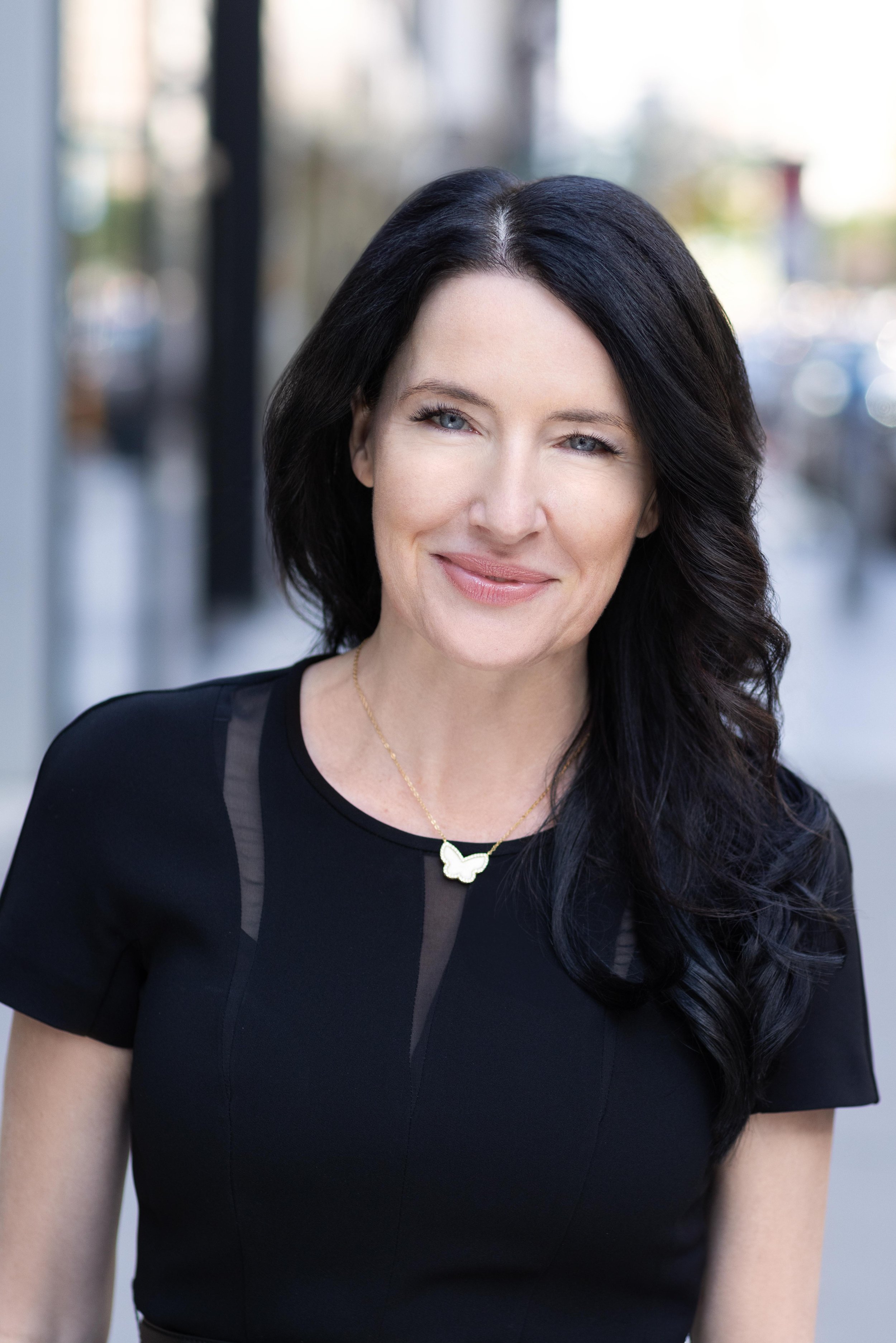 A woman with dark wavy hair smiling outdoors, wearing a black top with lace details and a butterfly necklace.