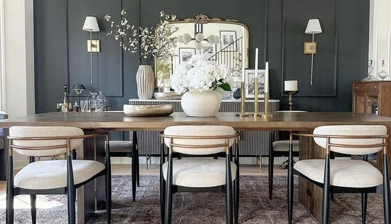 Modern dining room with dark wall panels, a large wooden table, six upholstered chairs, a mirror, vases, candles, and wall-mounted lamps.