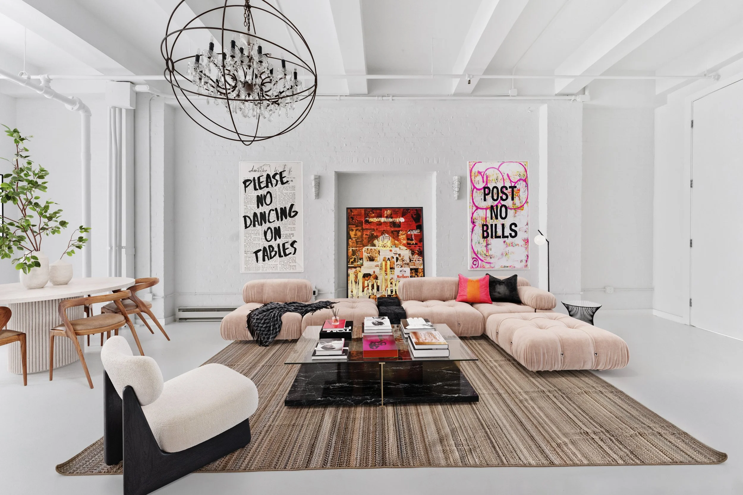 Modern living room with a blush pink sectional sofa, black marble coffee table, white walls, colorful artwork and posters, a chandelier, and a white column with green plants.